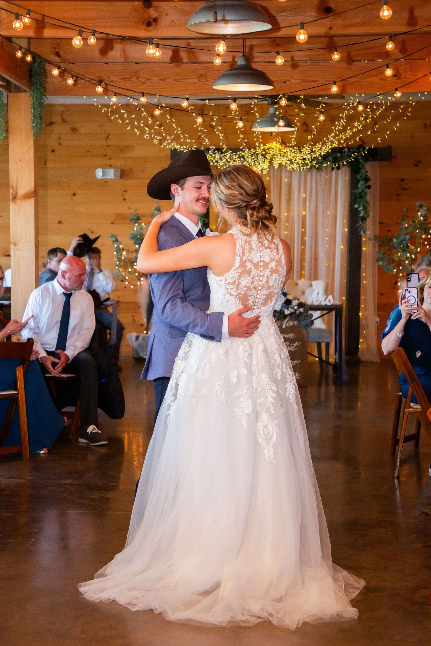 Bride and groom share their first dance in a rustic barn wedding venue adorned with warm string lights and greenery.