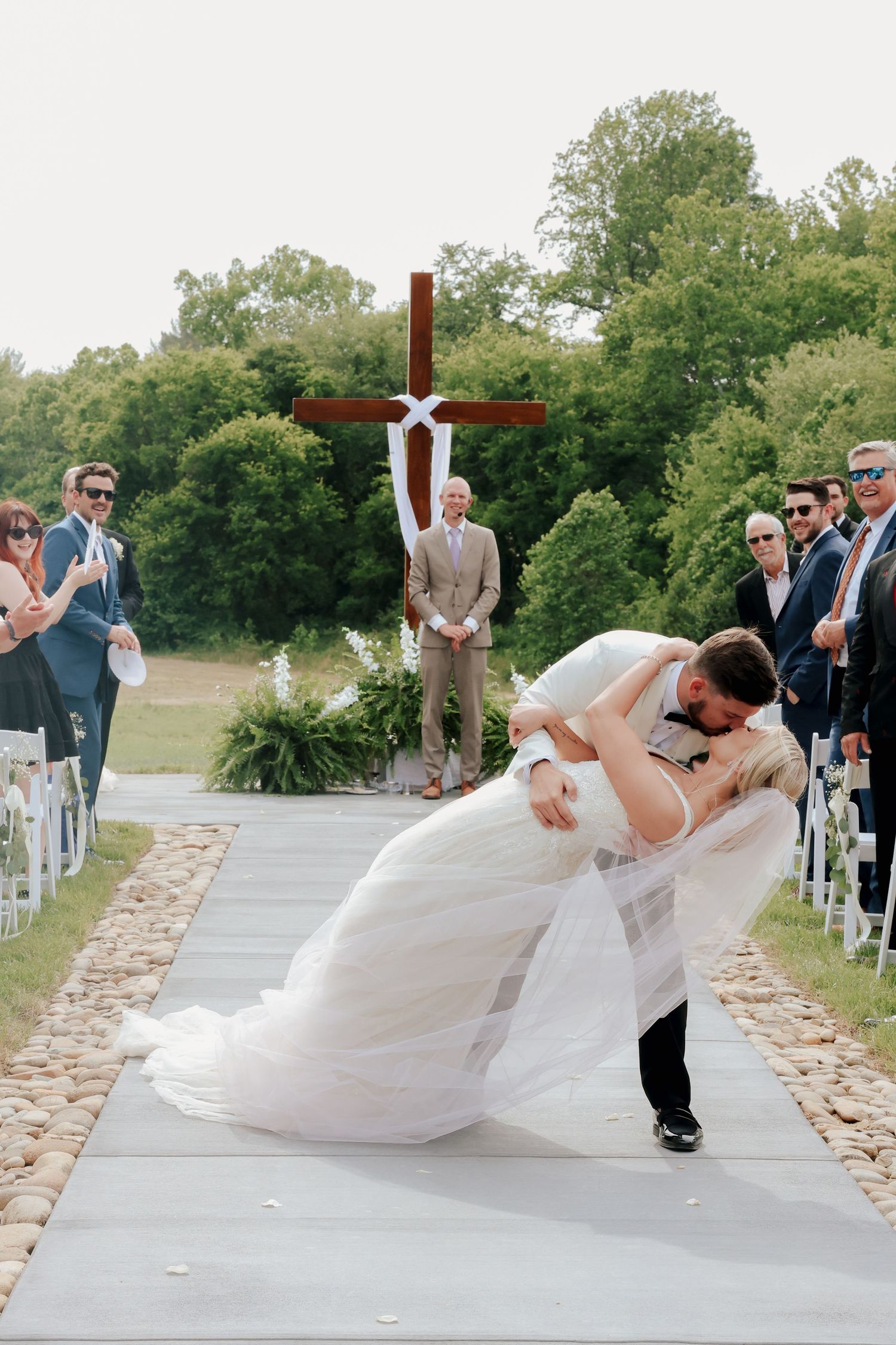 Groom dips bride in romantic kiss while guests cheer along aisle after outdoor wedding ceremony.