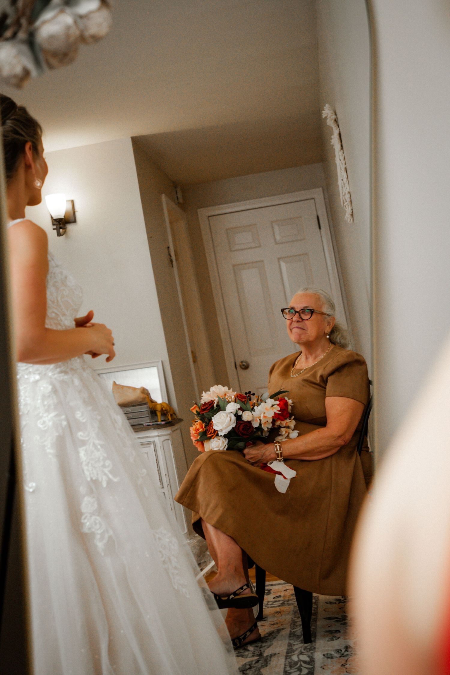 Elderly woman in brown dress holding bouquet watches bride in white lace gown during wedding preparations.