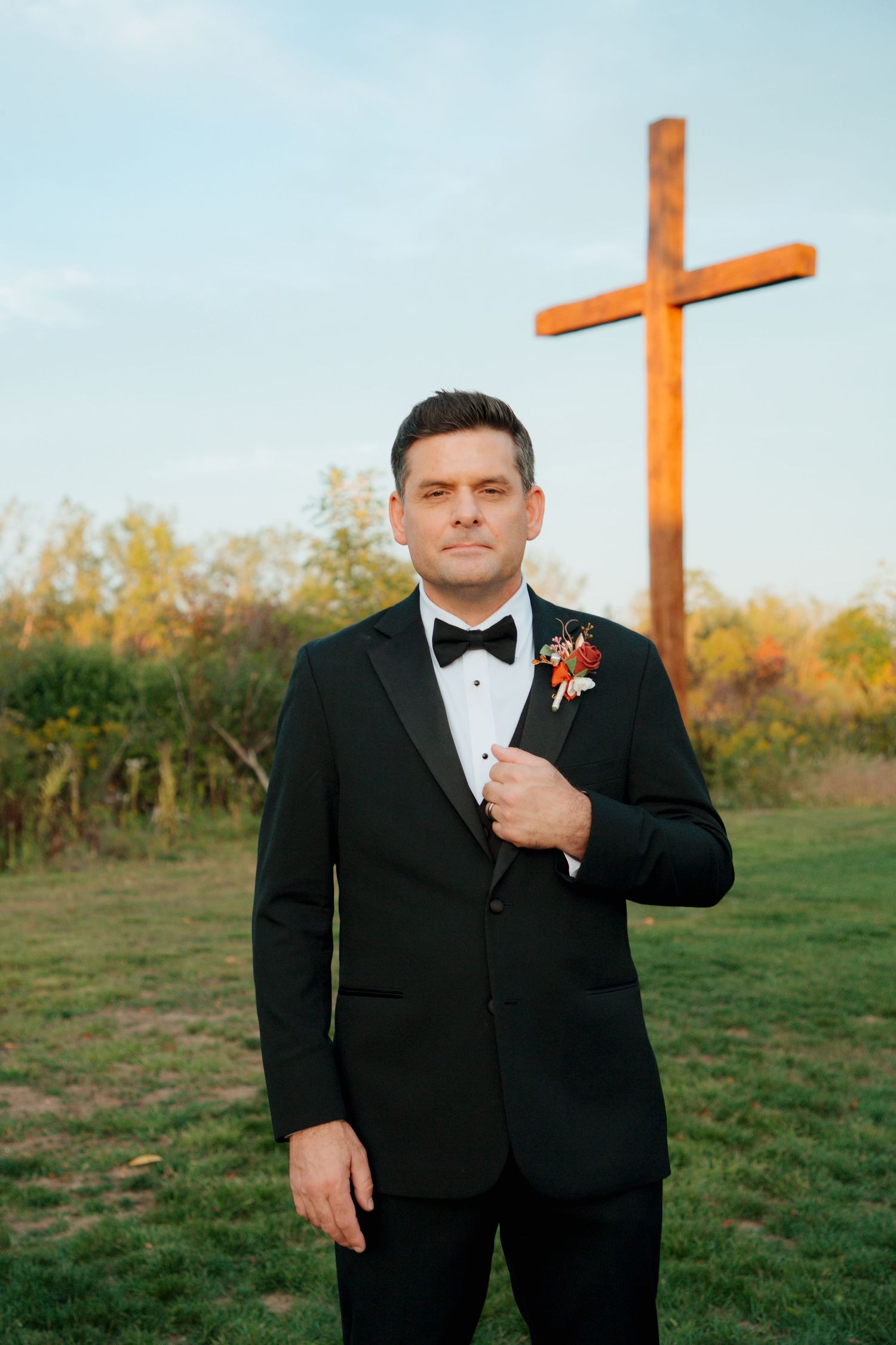 Groom in black tuxedo with floral boutonniere stands before a wooden cross outdoors.