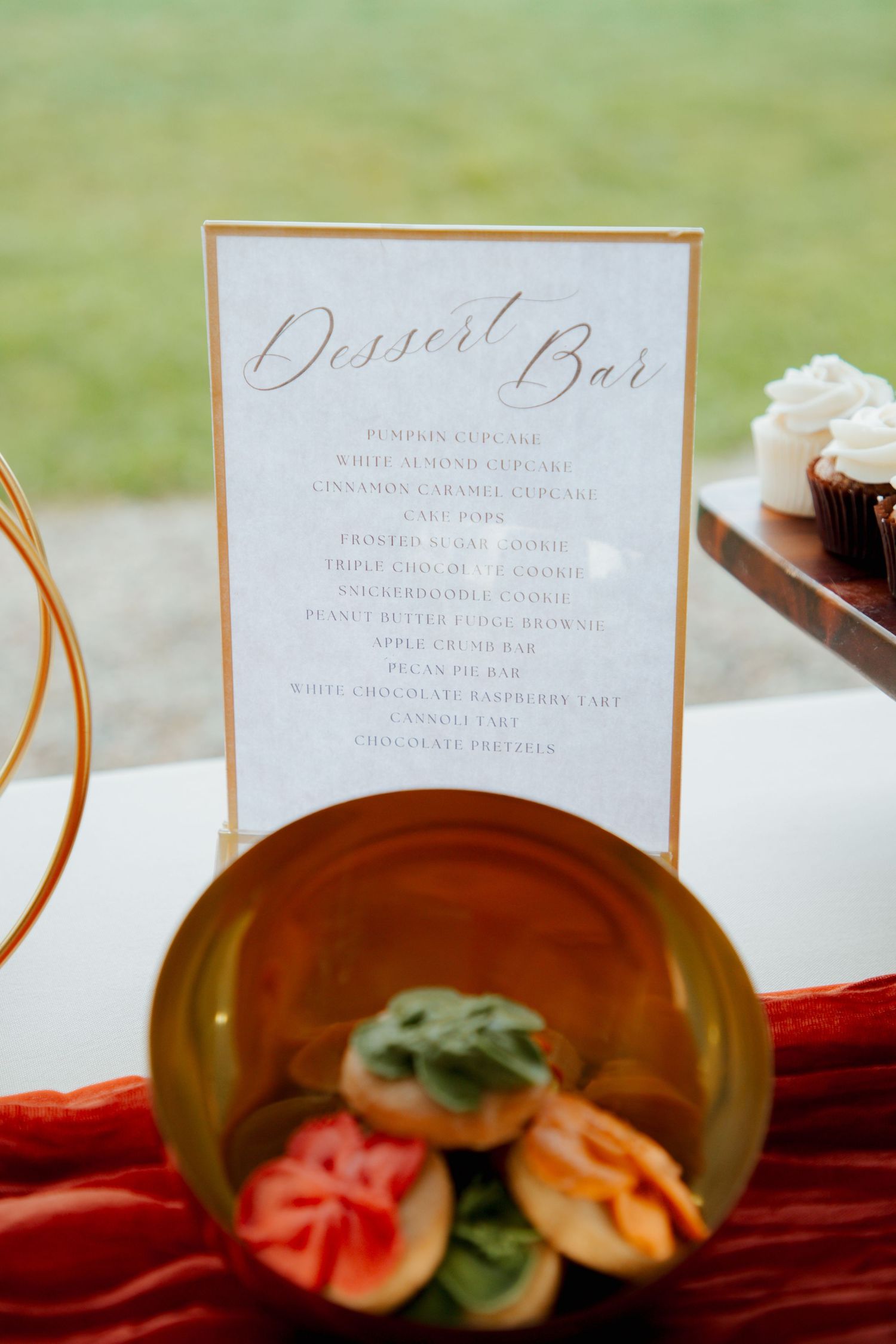 Wedding dessert bar menu sign in gold frame displaying cupcakes, cookies, and bars beside a gold plate with fruit.