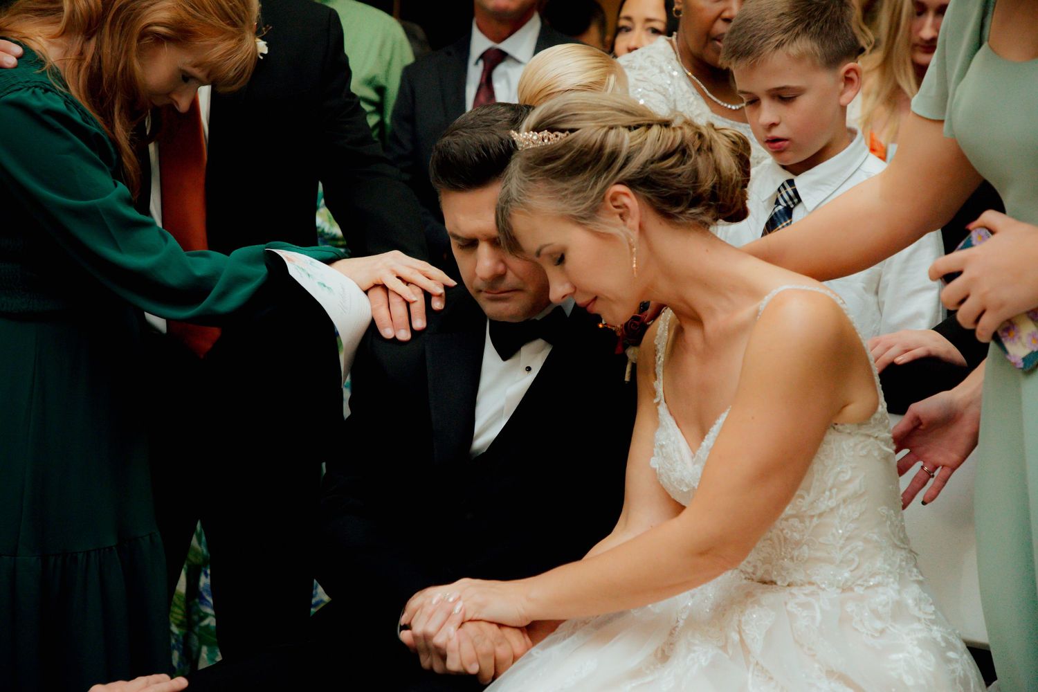 Bride and groom bow heads as wedding party members lay hands on them in prayer.
