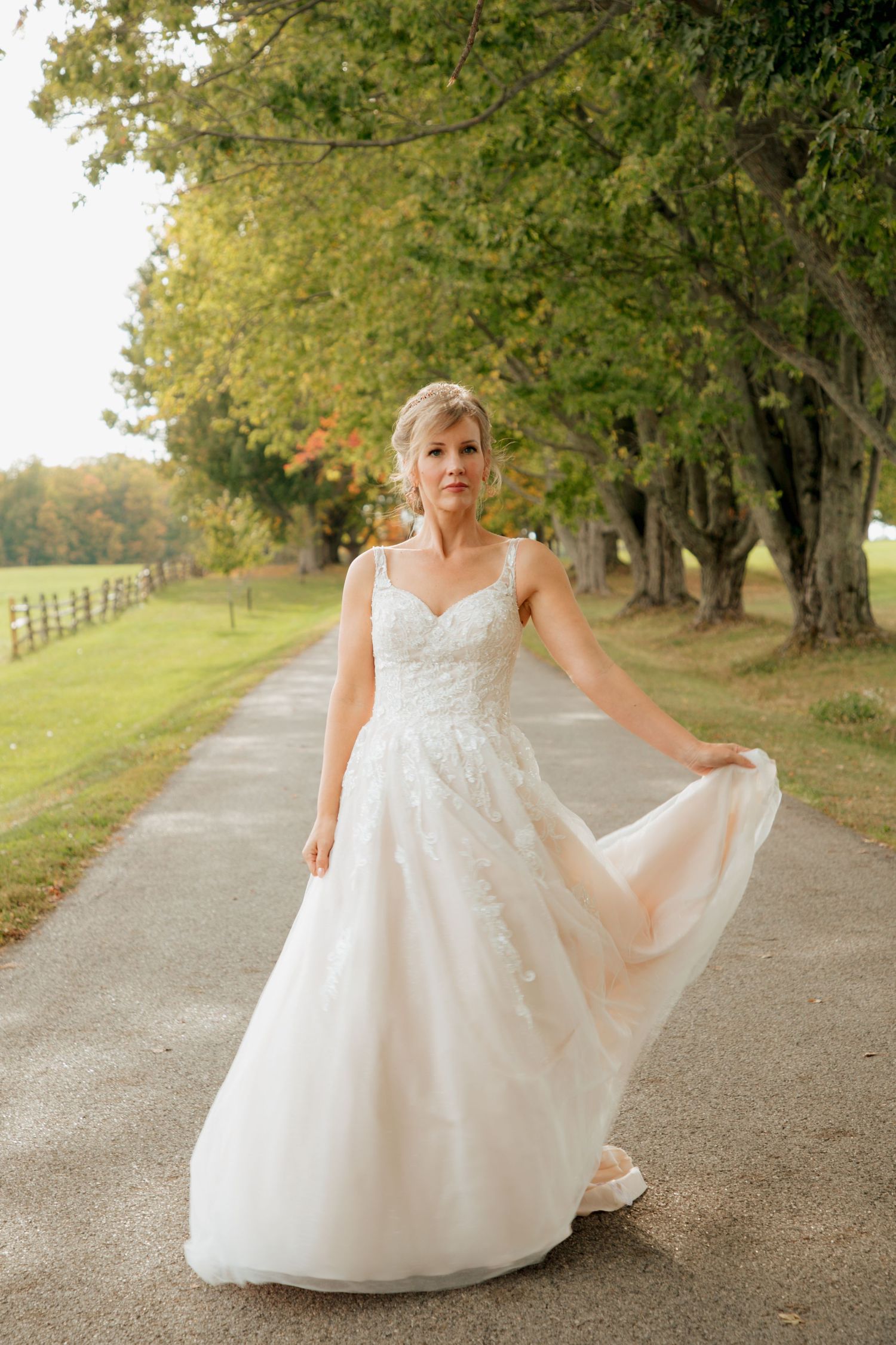 Bride in a lace A-line wedding gown gracefully twirling on a tree-lined path in autumn.