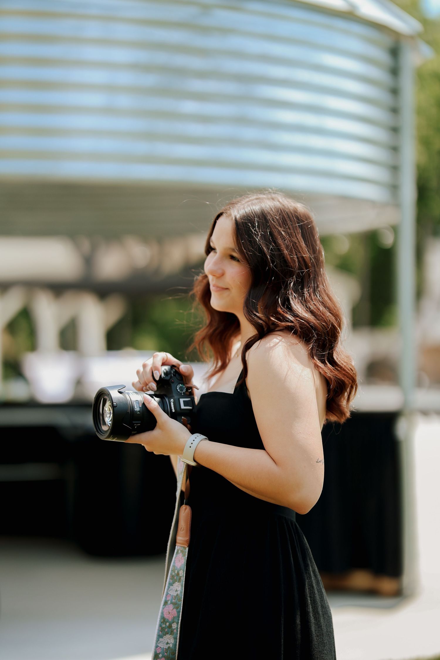 Female photographer in black dress holds camera with floral strap while looking away outdoors.