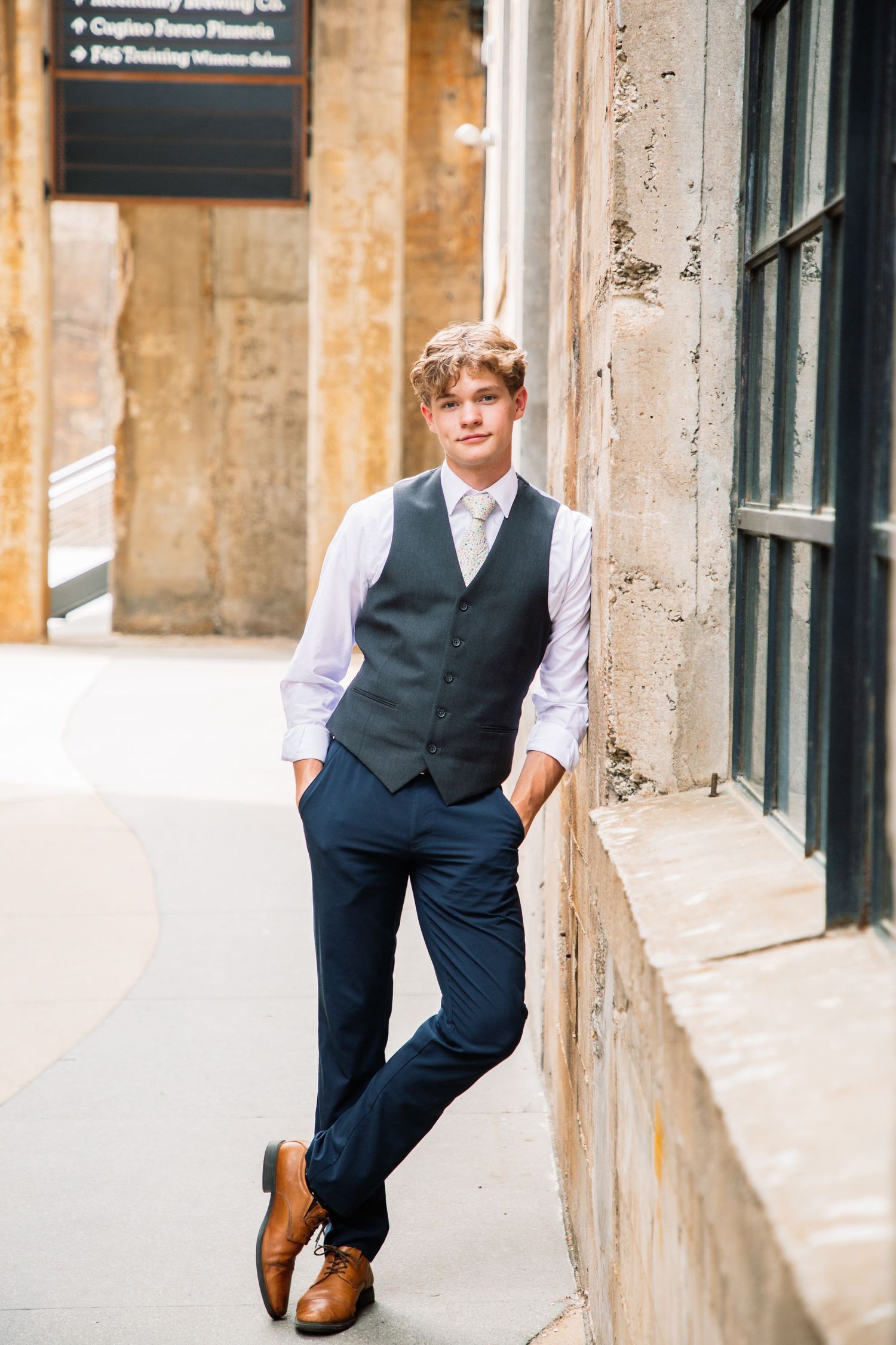 Young man in navy vest white shirt and tie leaning against stone building wall outdoors.