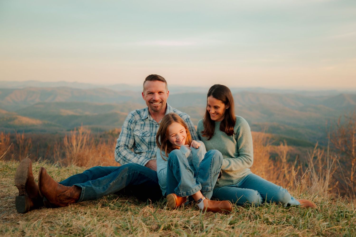 A laughing family of three relaxes on a grassy hillside with breathtaking autumn mountain views at sunset.