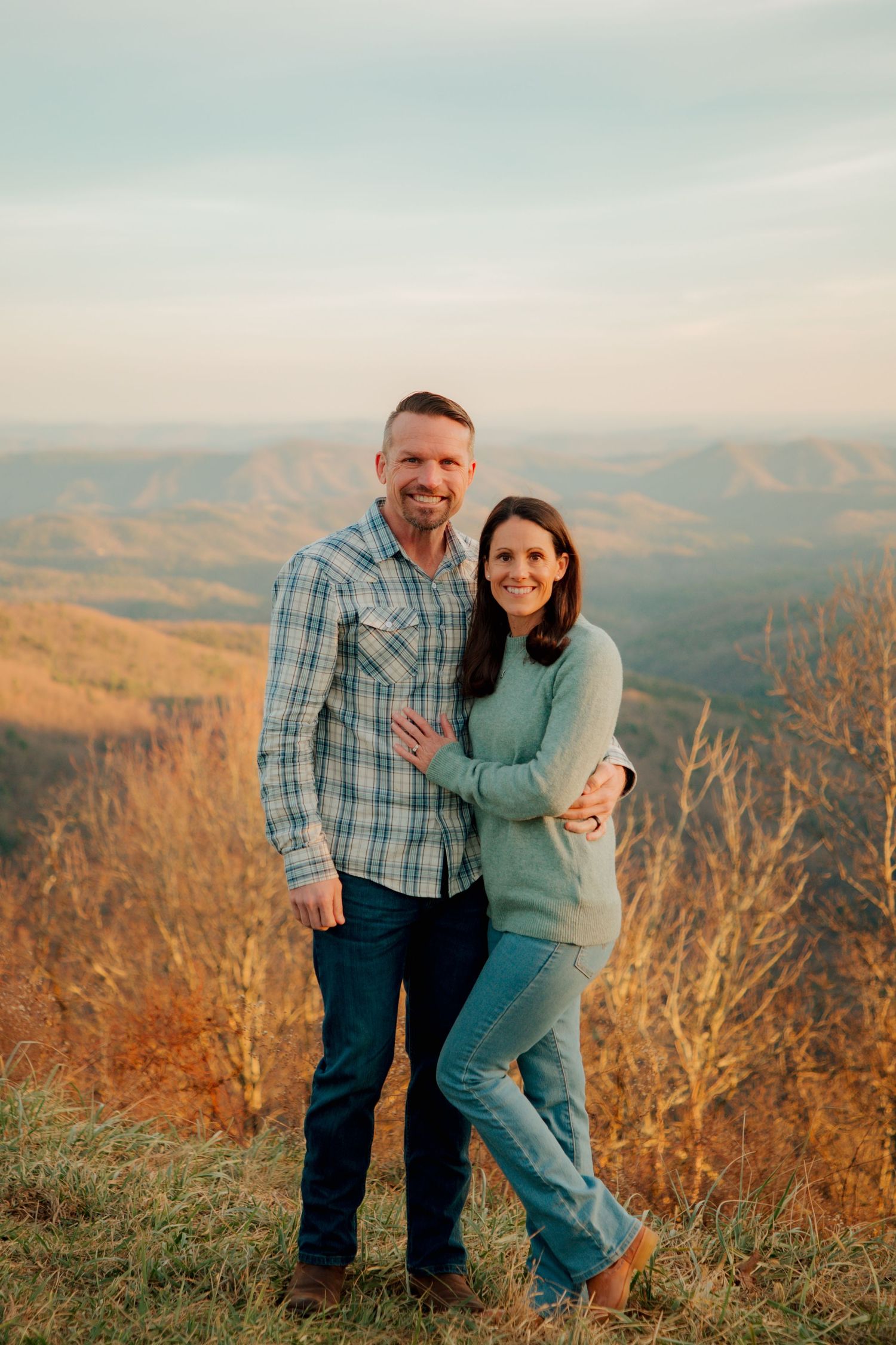 Smiling couple embraces while standing on a hilltop with breathtaking autumn mountain views.