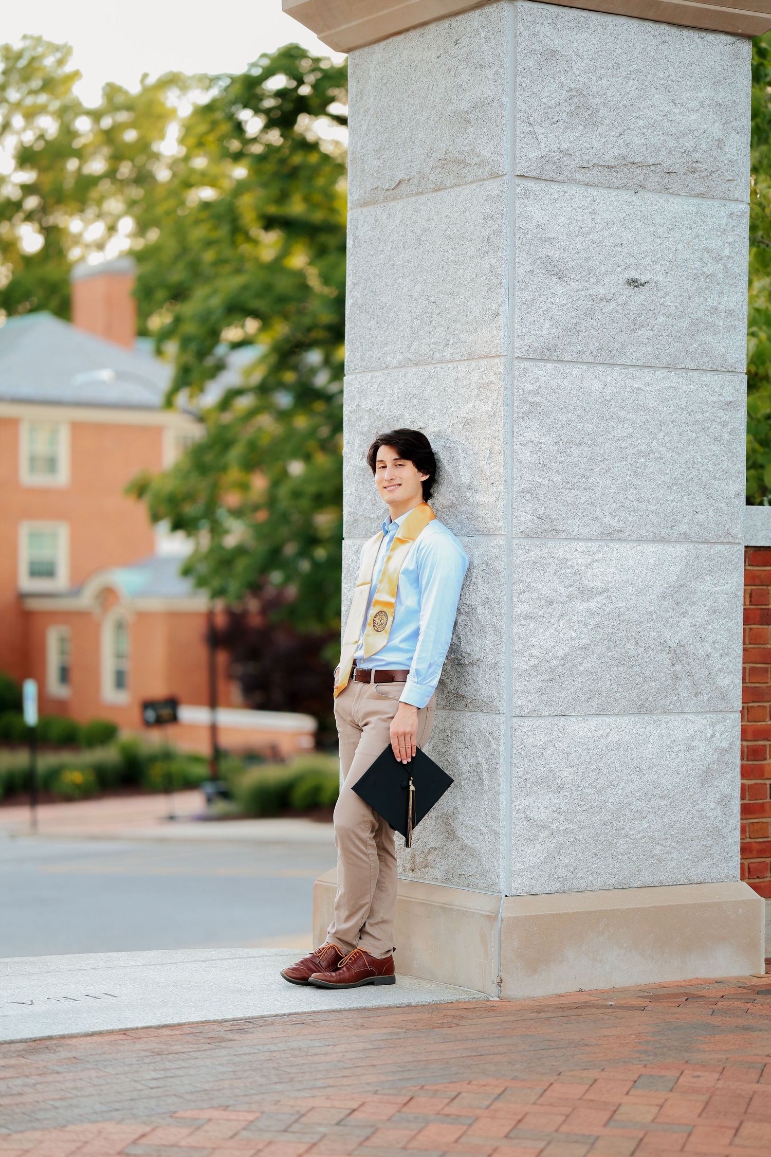 Young male graduate holding cap leans against stone entrance pillar on brick university campus.