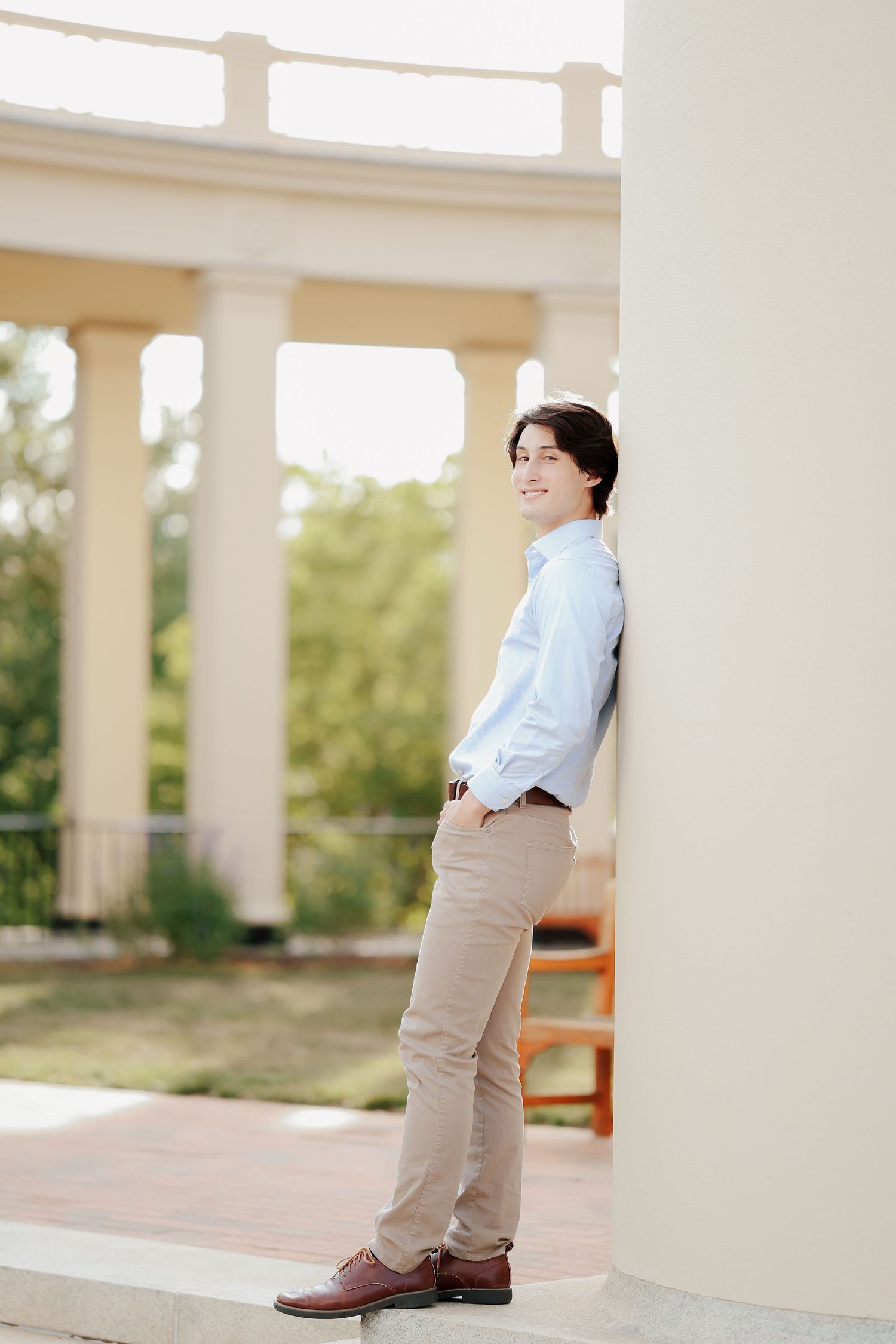 Smiling young man in blue shirt leans against a white column at an outdoor campus colonnade.