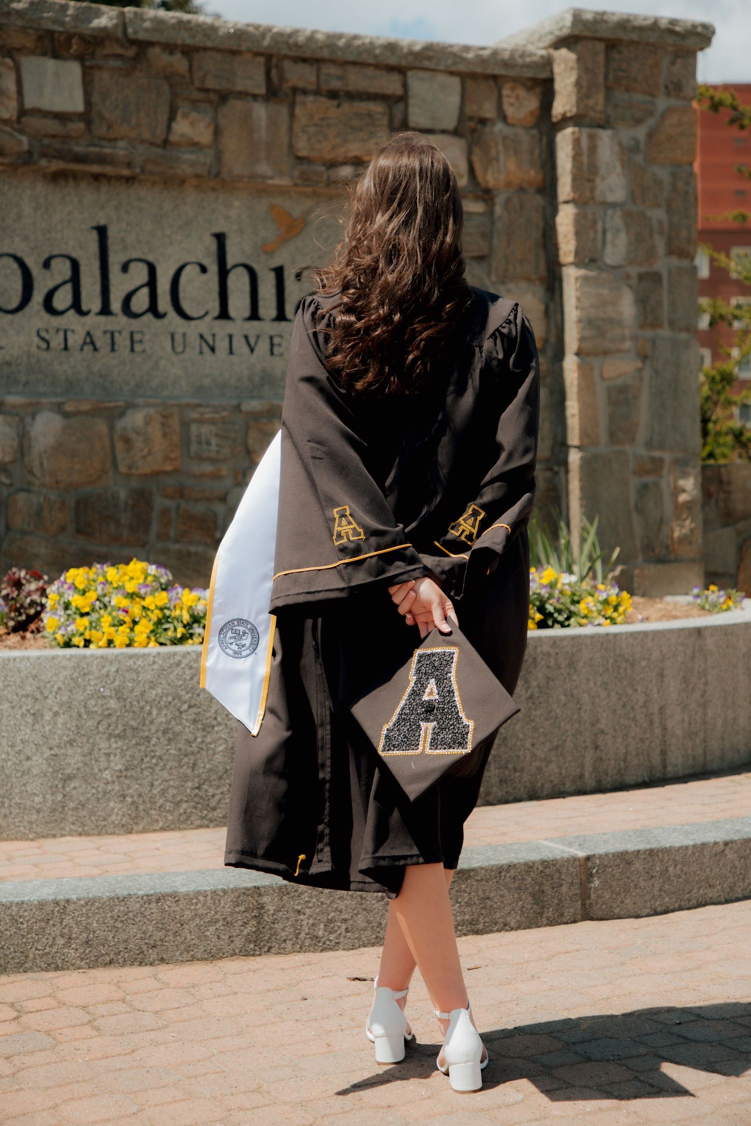 Graduate in black gown holding decorated cap with letter A before Appalachian State University sign.