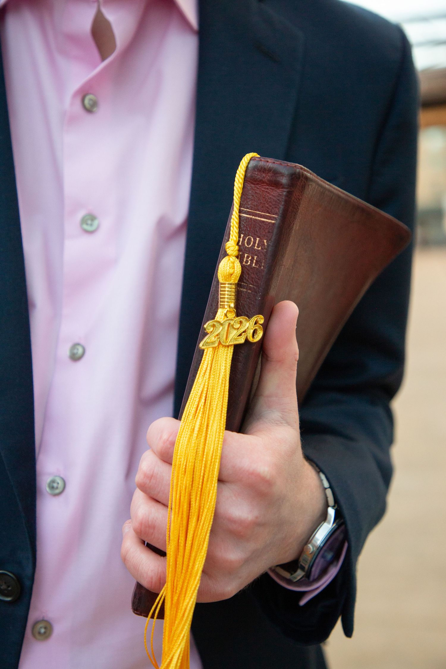 Close-up of hands holding Holy Bible with gold 2026 graduation tassel against blue suit jacket.