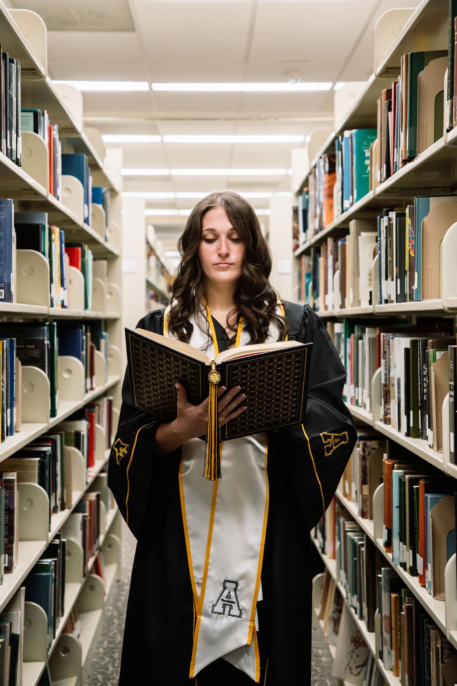 Female graduate in black robe reading book between library shelves.
