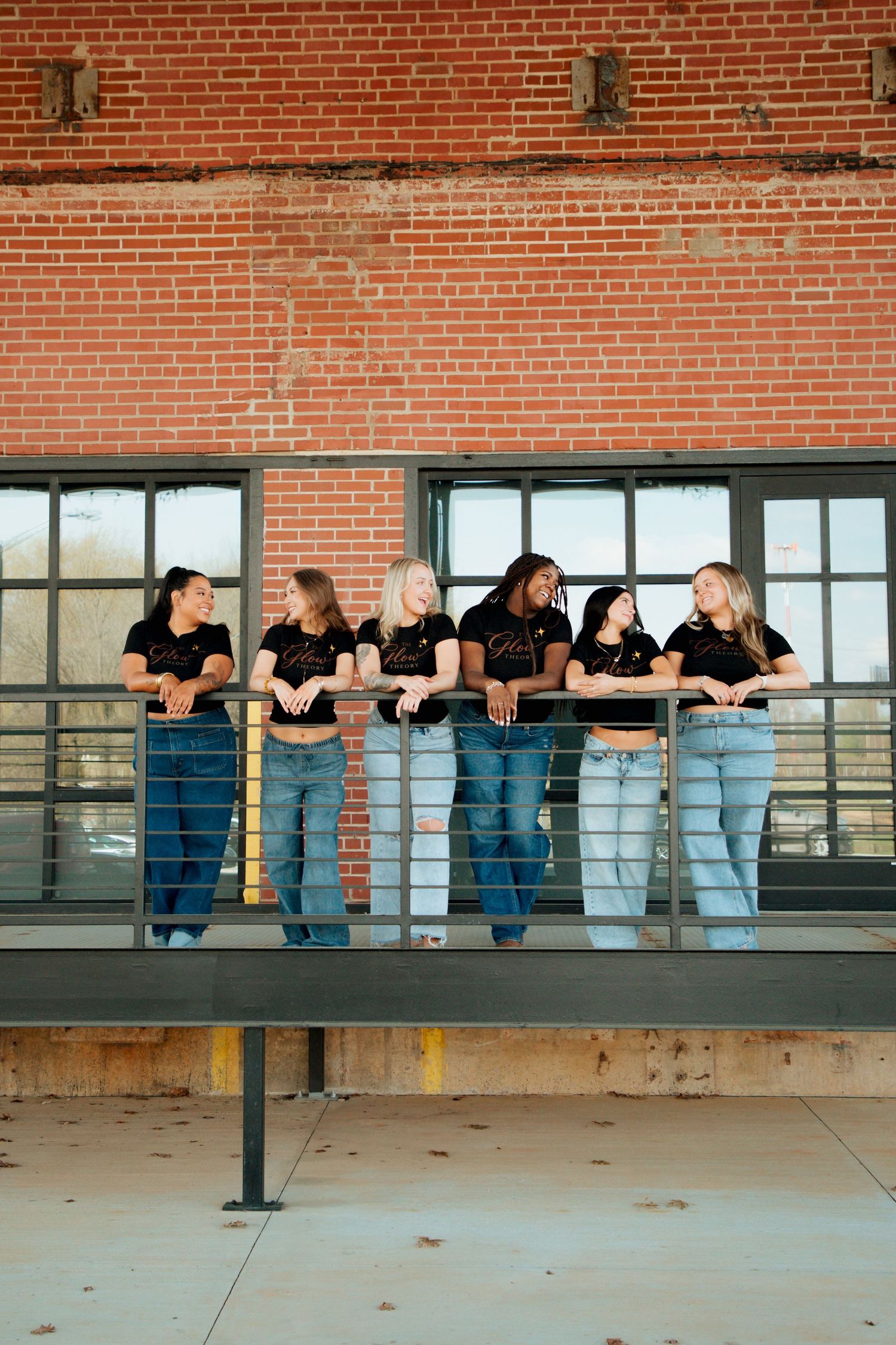 Six young women in matching black tops and jeans pose together leaning on a metal railing in front of a brick building.