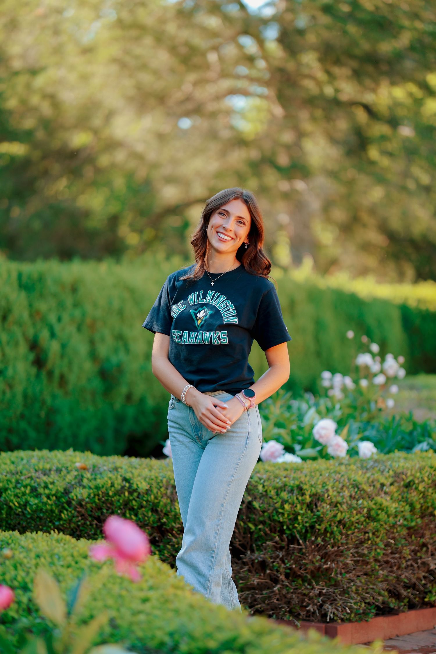 Smiling young woman in dark college t-shirt and jeans standing in manicured garden with flowers.