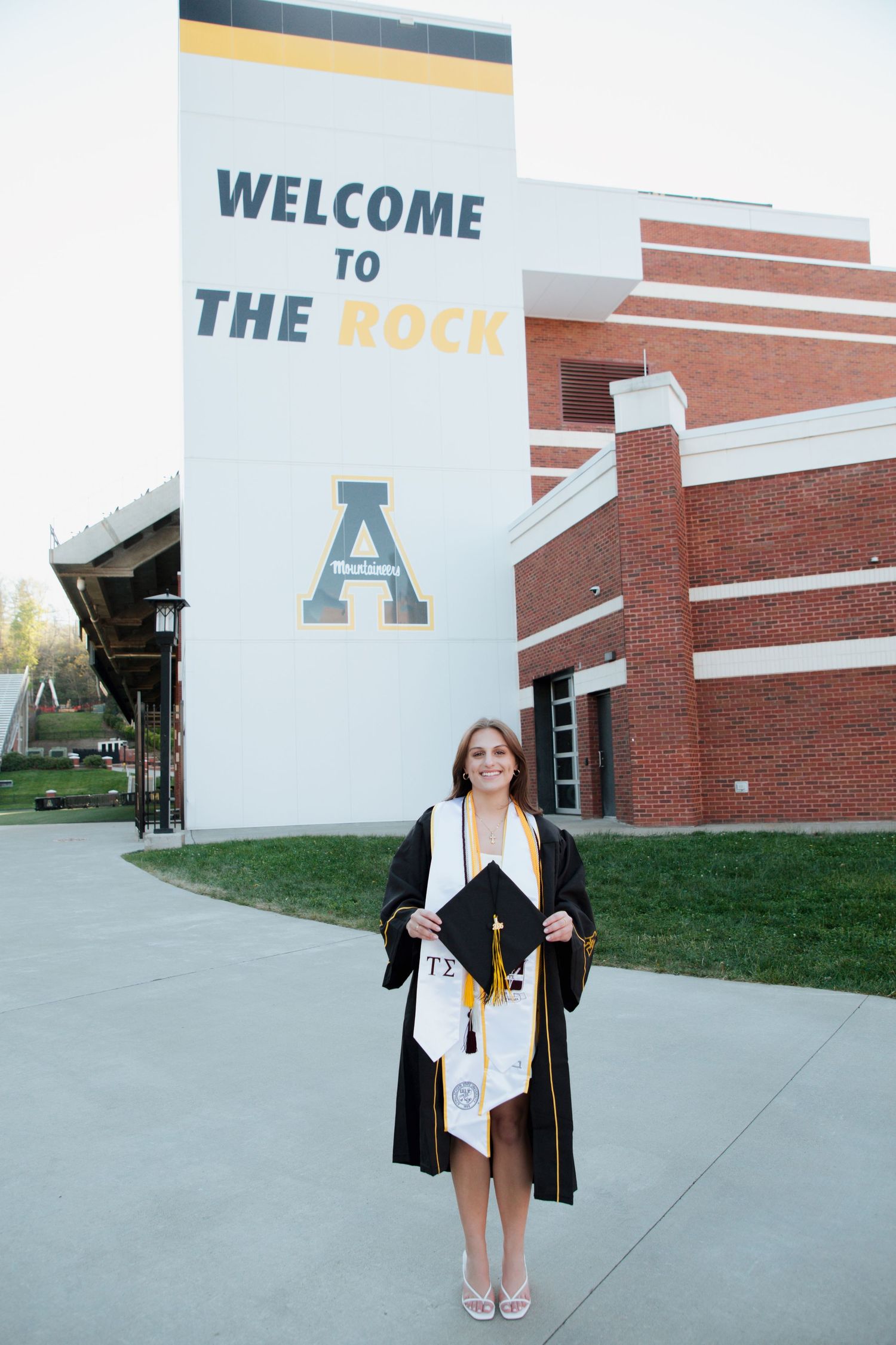 Smiling graduate holding cap in front of Welcome to The Rock Appalachian State stadium sign.