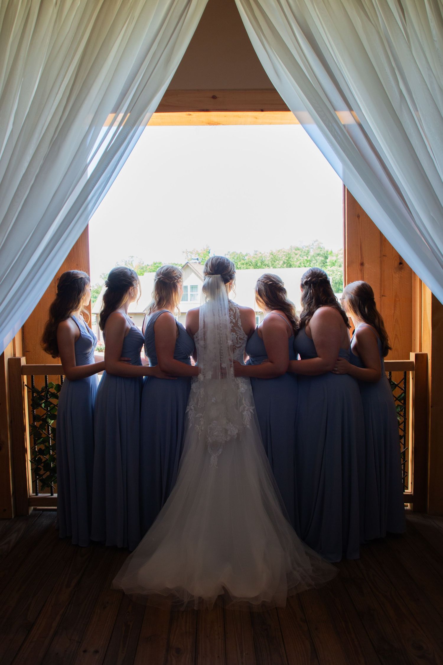Bride and five bridesmaids in dusty blue dresses stand with backs to camera framed by white drapes.