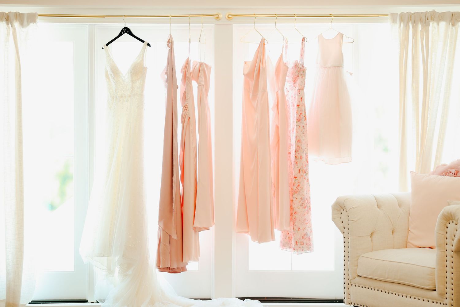 White wedding gown and blush pink bridesmaid dresses hanging on a rack near bright windows.