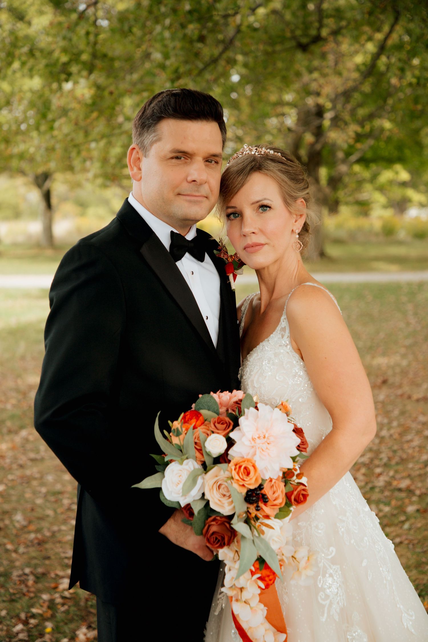 Bride and groom pose together in autumn park with fall bouquet of orange and pink roses.