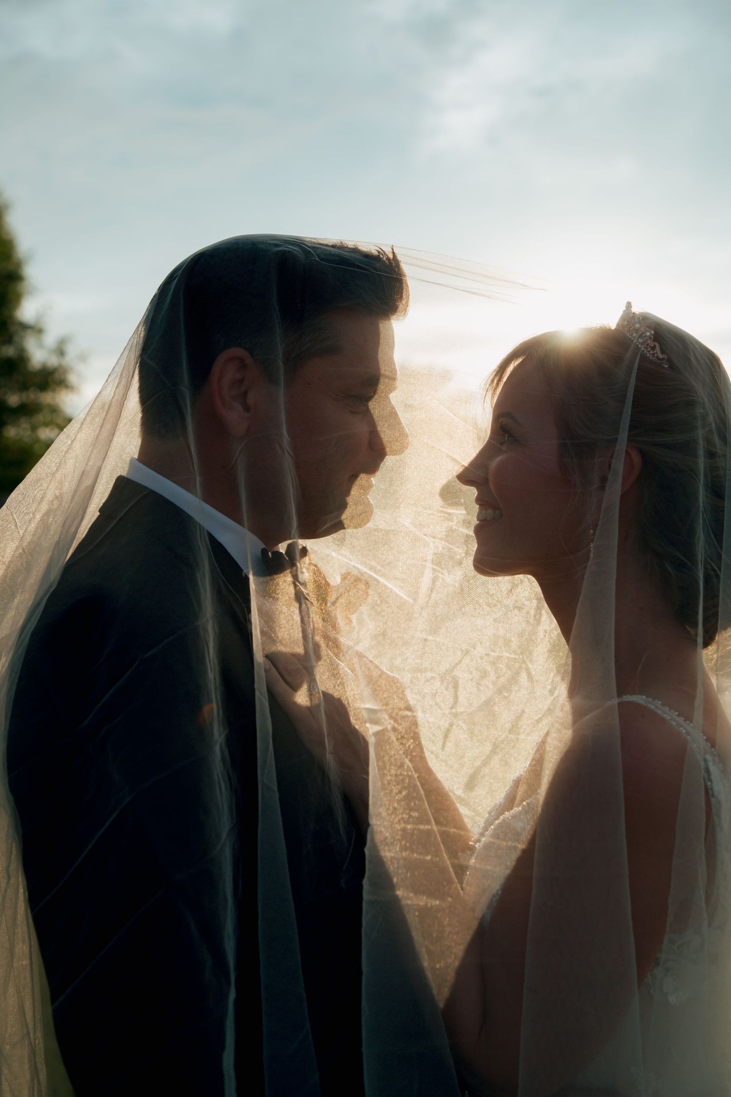 Bride and groom share an intimate moment under the wedding veil with stunning golden sunlight glowing behind them.