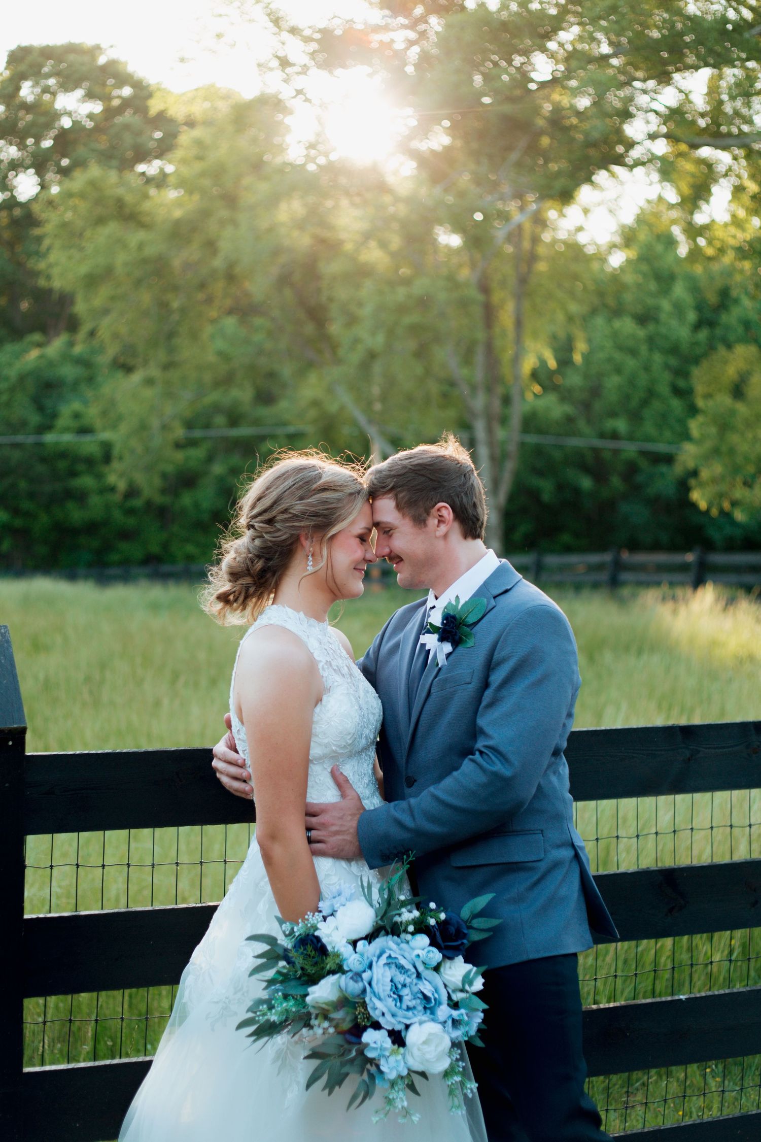 Bride and groom touch foreheads romantically by rustic fence at golden hour.