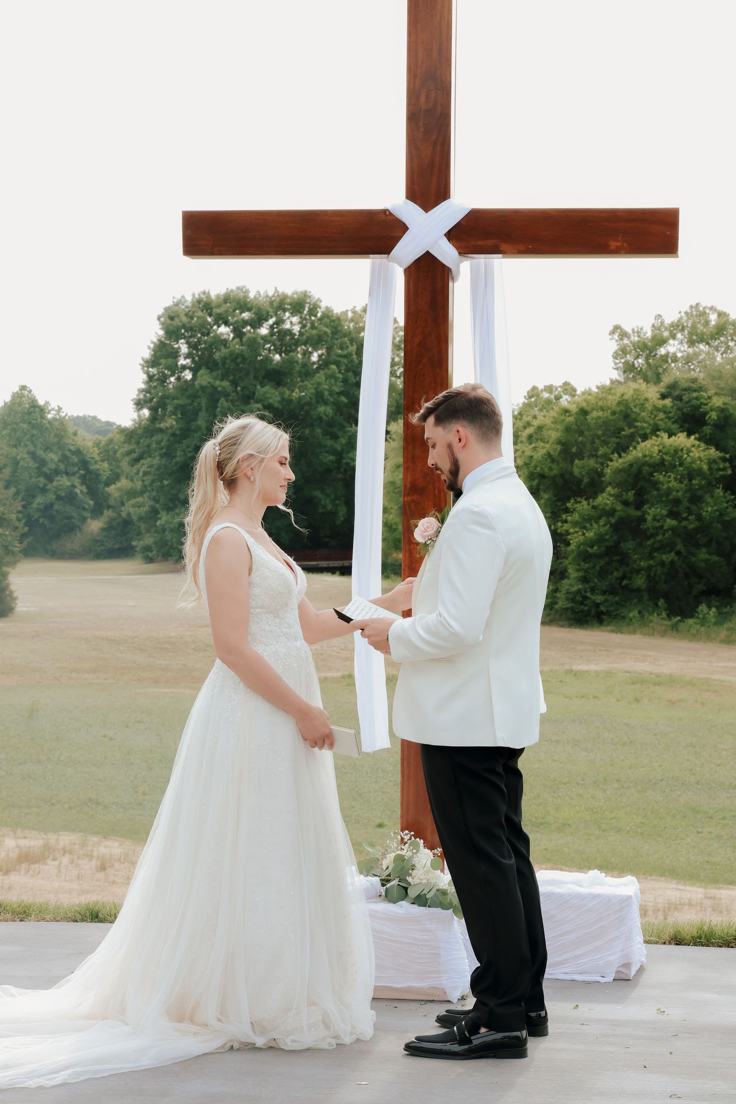 Groom in white jacket reading vows to bride in white gown during outdoor wedding ceremony before large wooden cross.
