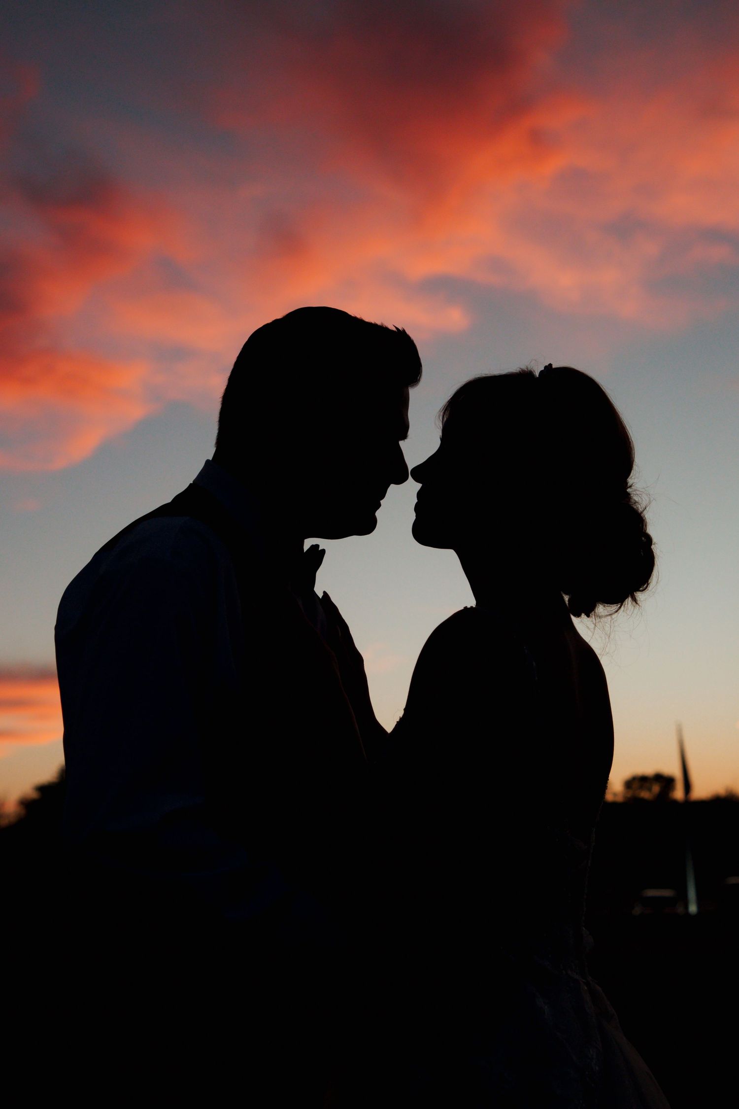Romantic silhouette of bride and groom nearly kissing against vibrant pink sunset sky.