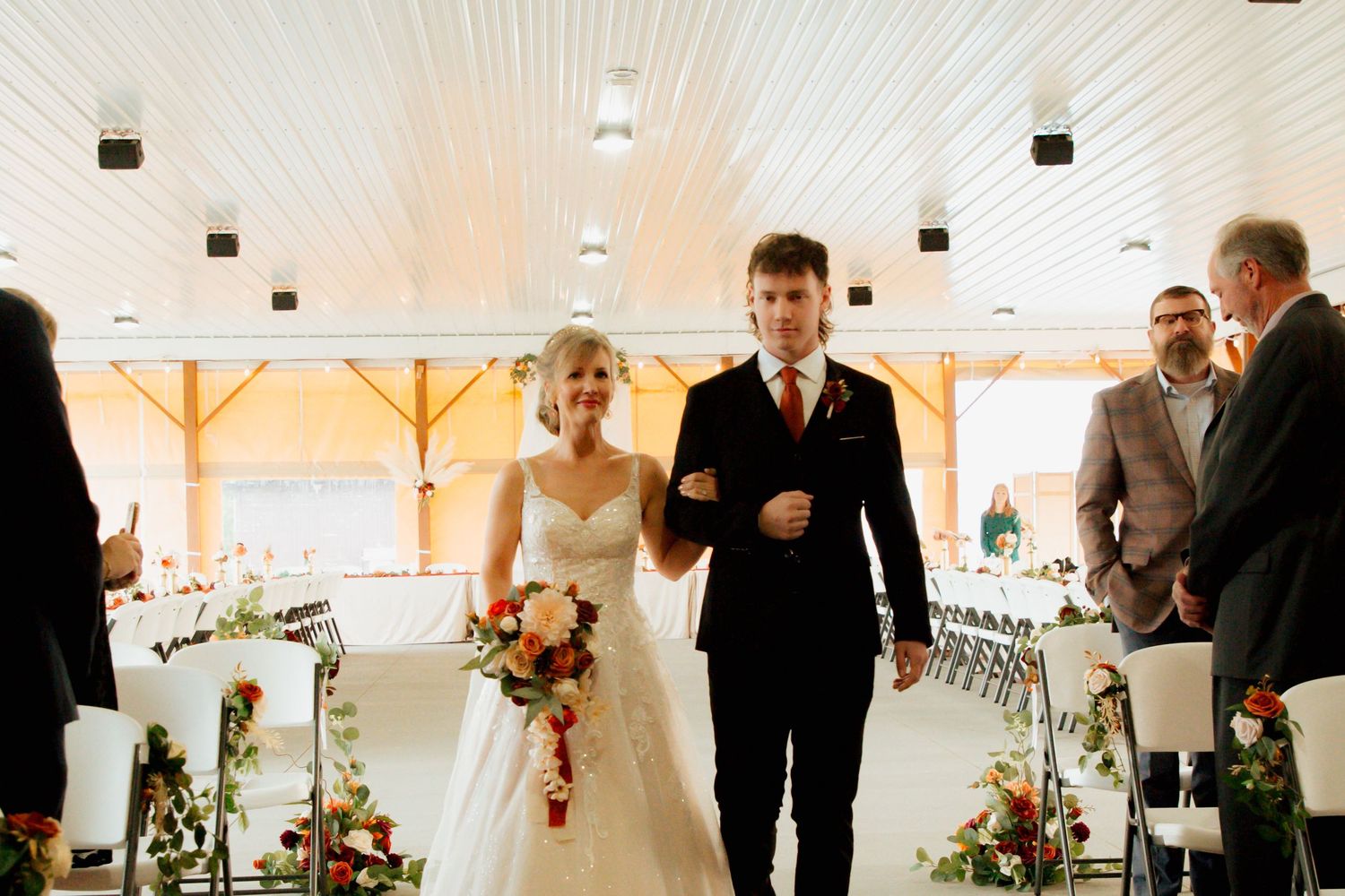 A smiling bride holding a bouquet walks arm-in-arm with her groom down a decorated wedding ceremony aisle.
