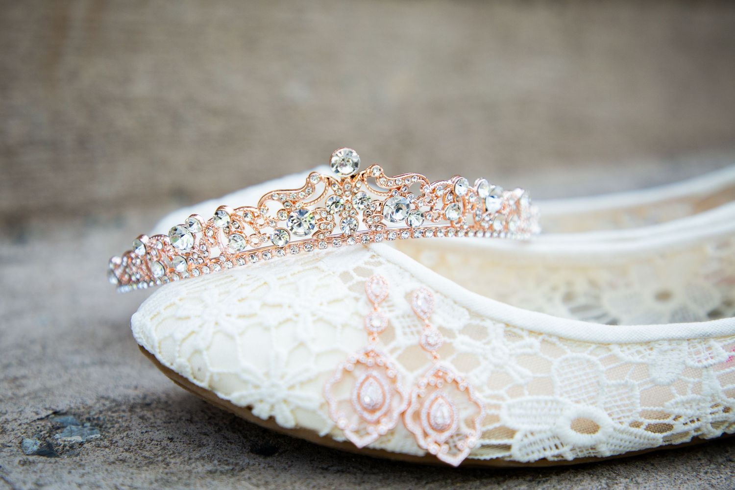 Rose gold rhinestone bridal tiara crown resting on white lace wedding flat shoes on a stone surface.