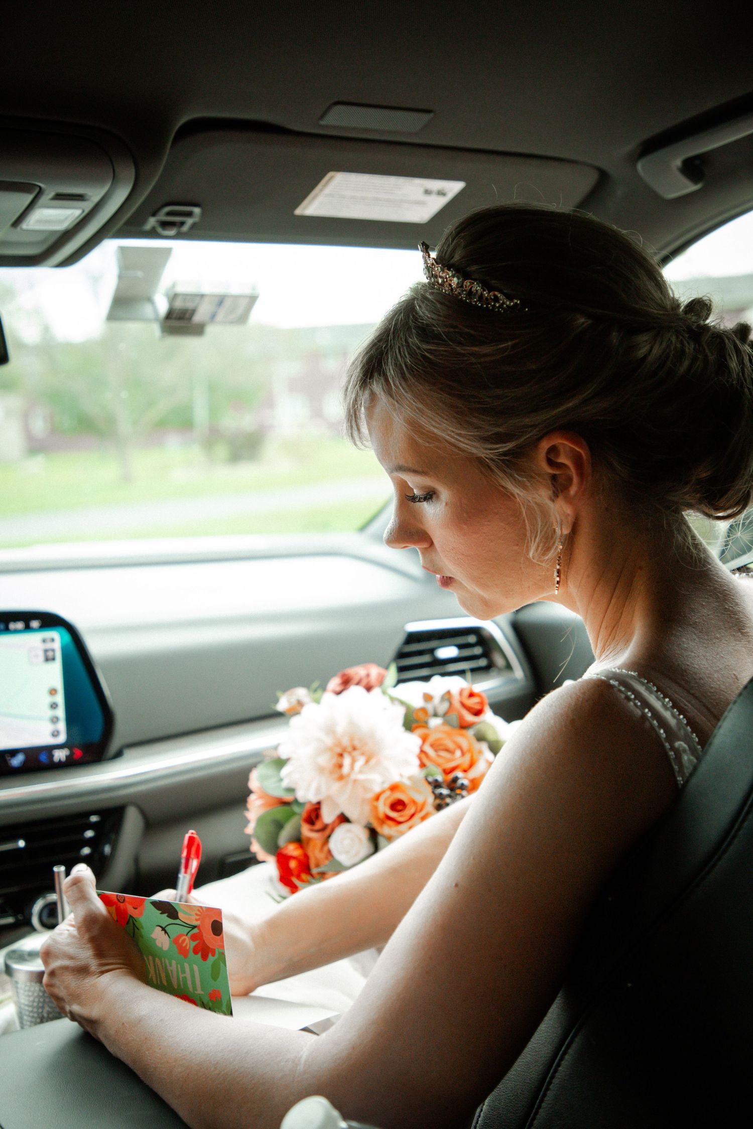 Bride in white gown with tiara reads a card while holding floral bouquet inside a vehicle.
