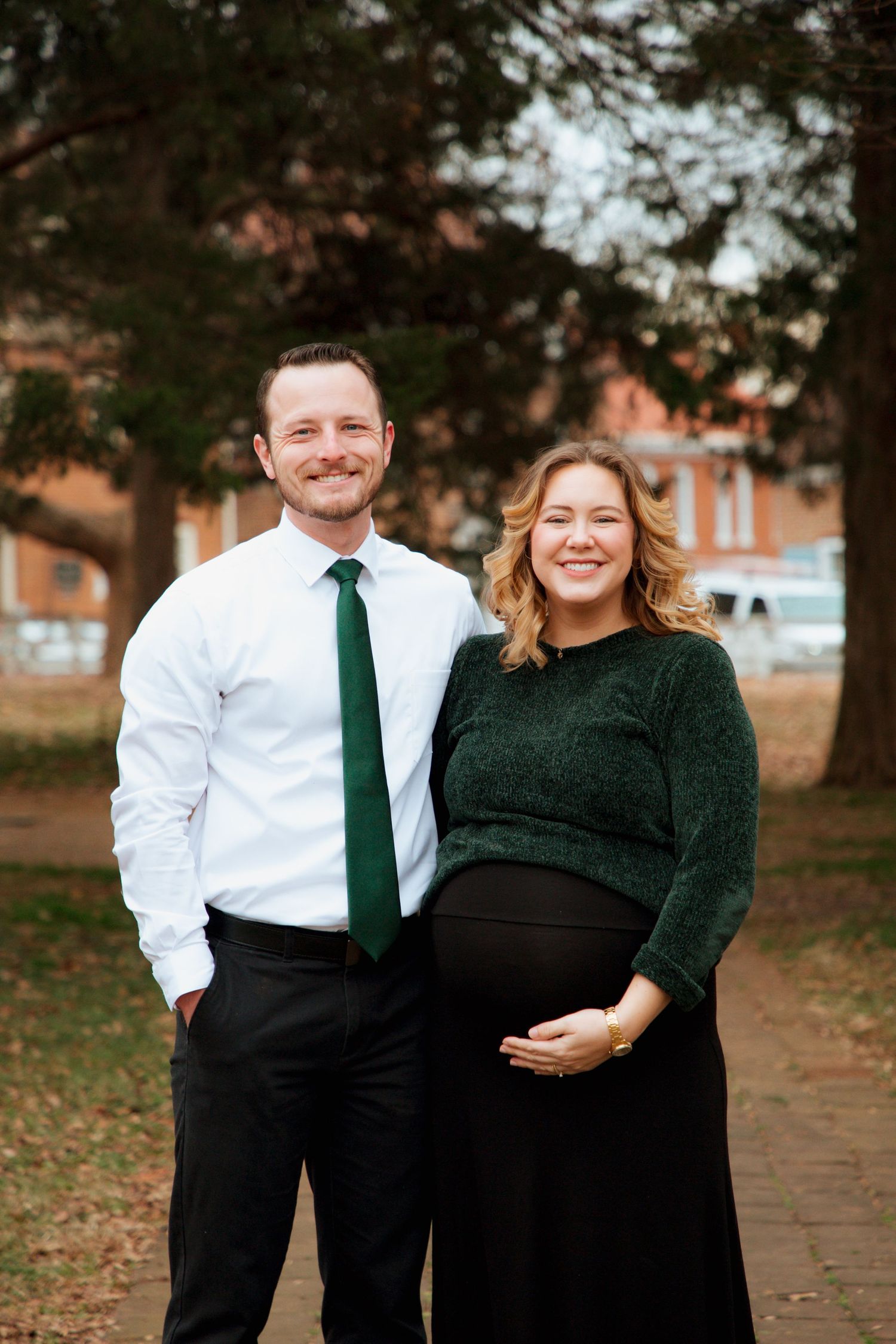 Smiling pregnant woman in green sweater and black skirt poses with man in white shirt and green tie in park.