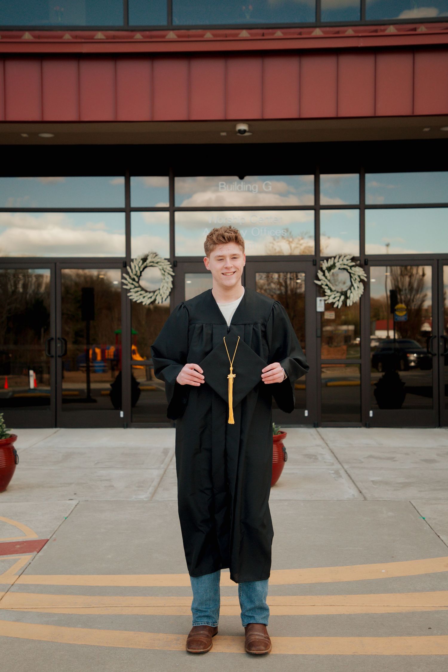 Young man in black graduation gown and gold tassel smiling in front of school entrance.