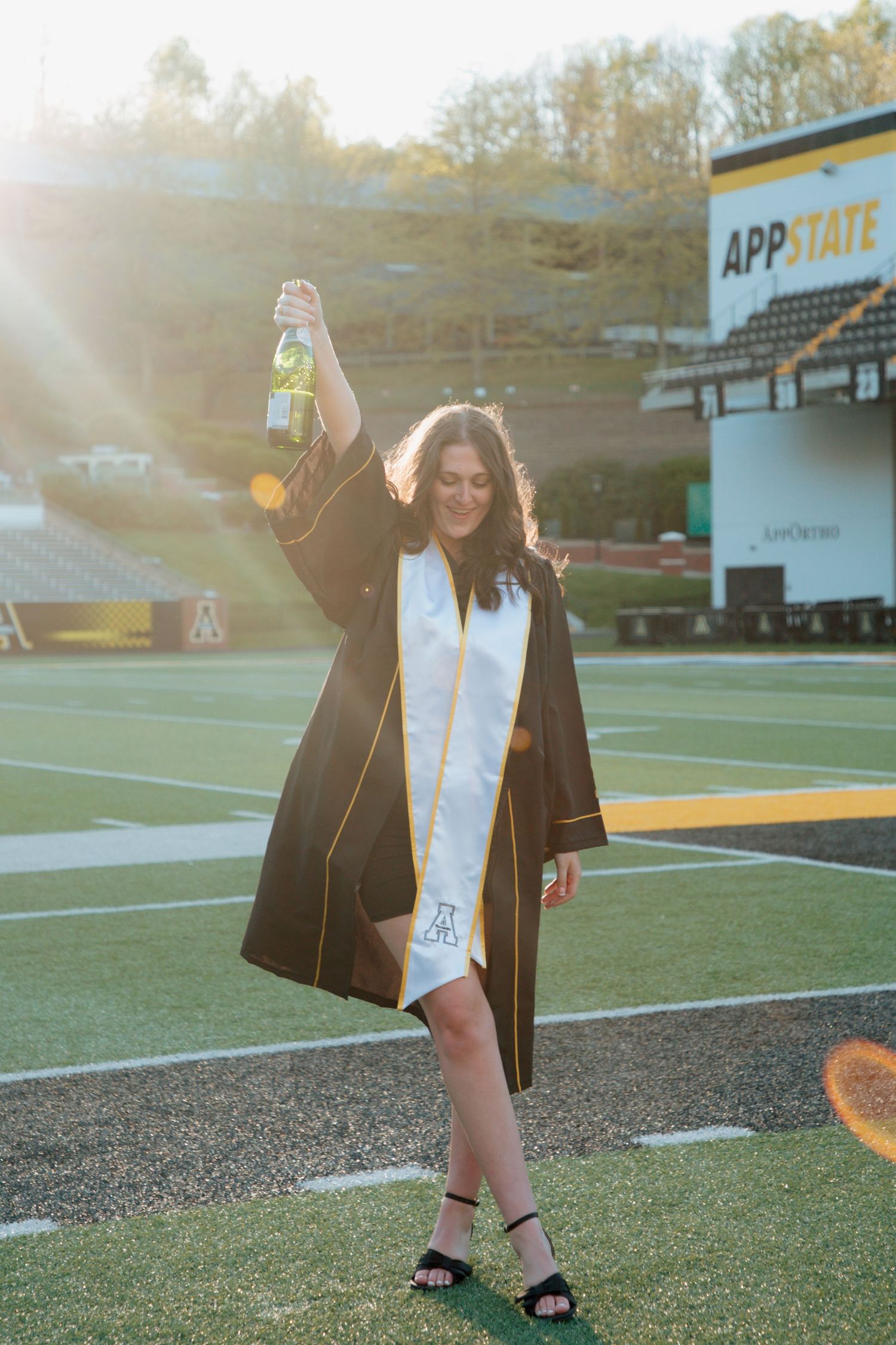 Graduate celebrates holding champagne bottle on App State football field in golden sunlight.