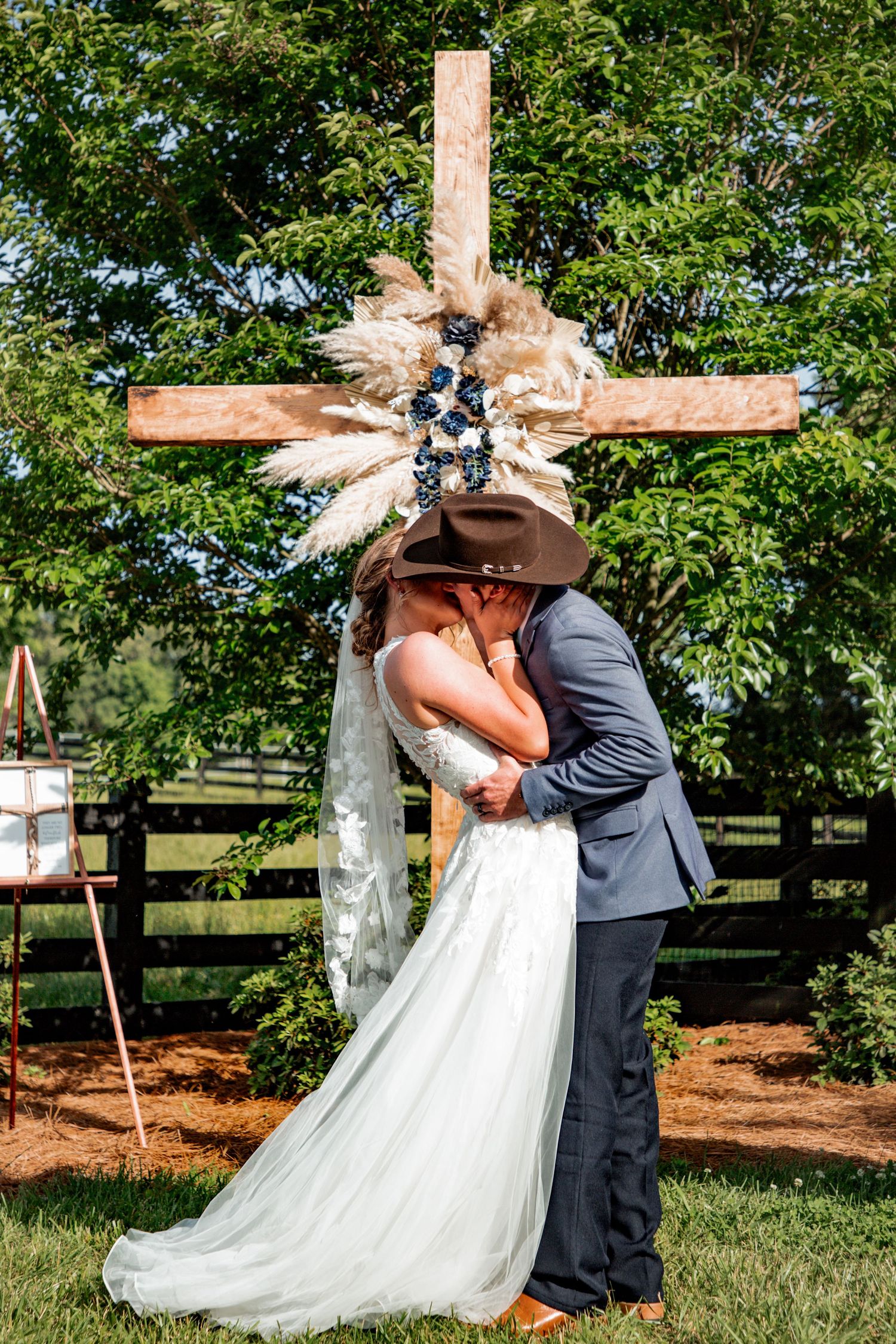Bride and groom share first kiss in front of rustic wooden cross decorated with pampas grass and blue flowers at outdoor wedding.