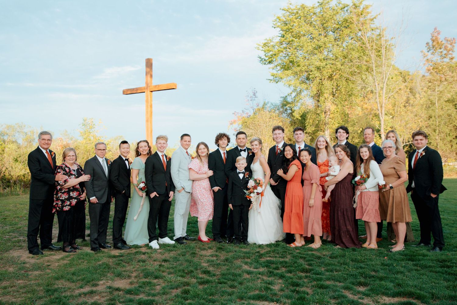 Large wedding party posing outdoors near a wooden cross on a sunny autumn day.