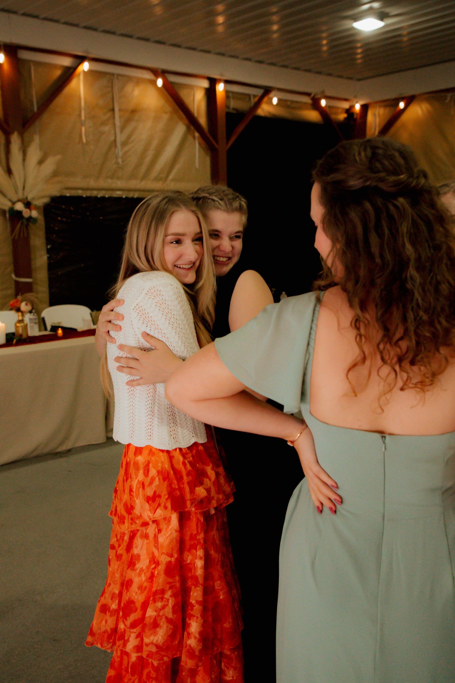 Three young women sharing a warm embrace at an elegant wedding reception venue.