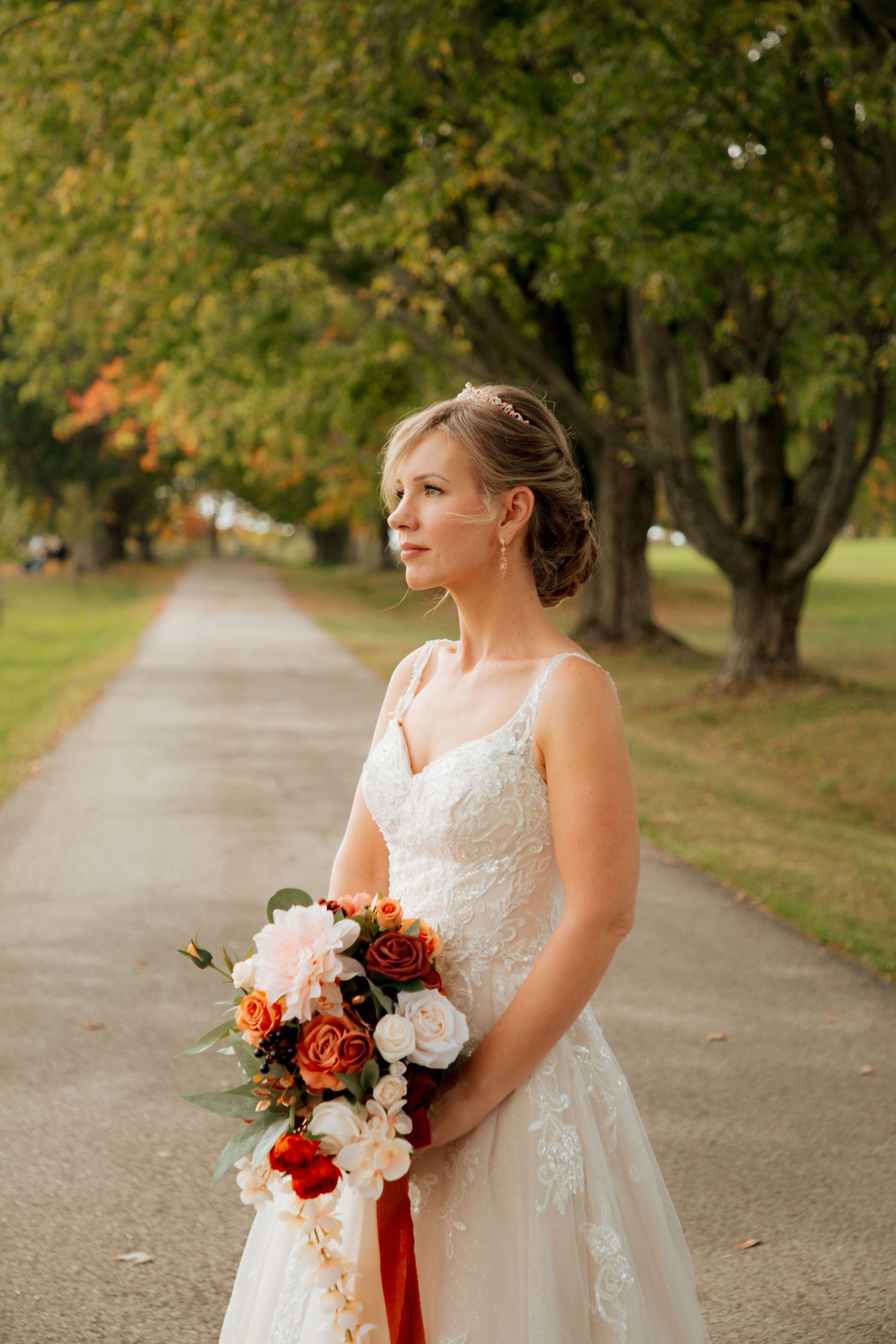 Bride in a lace wedding gown gazes thoughtfully while holding a cascading fall bouquet on a tree-lined path.
