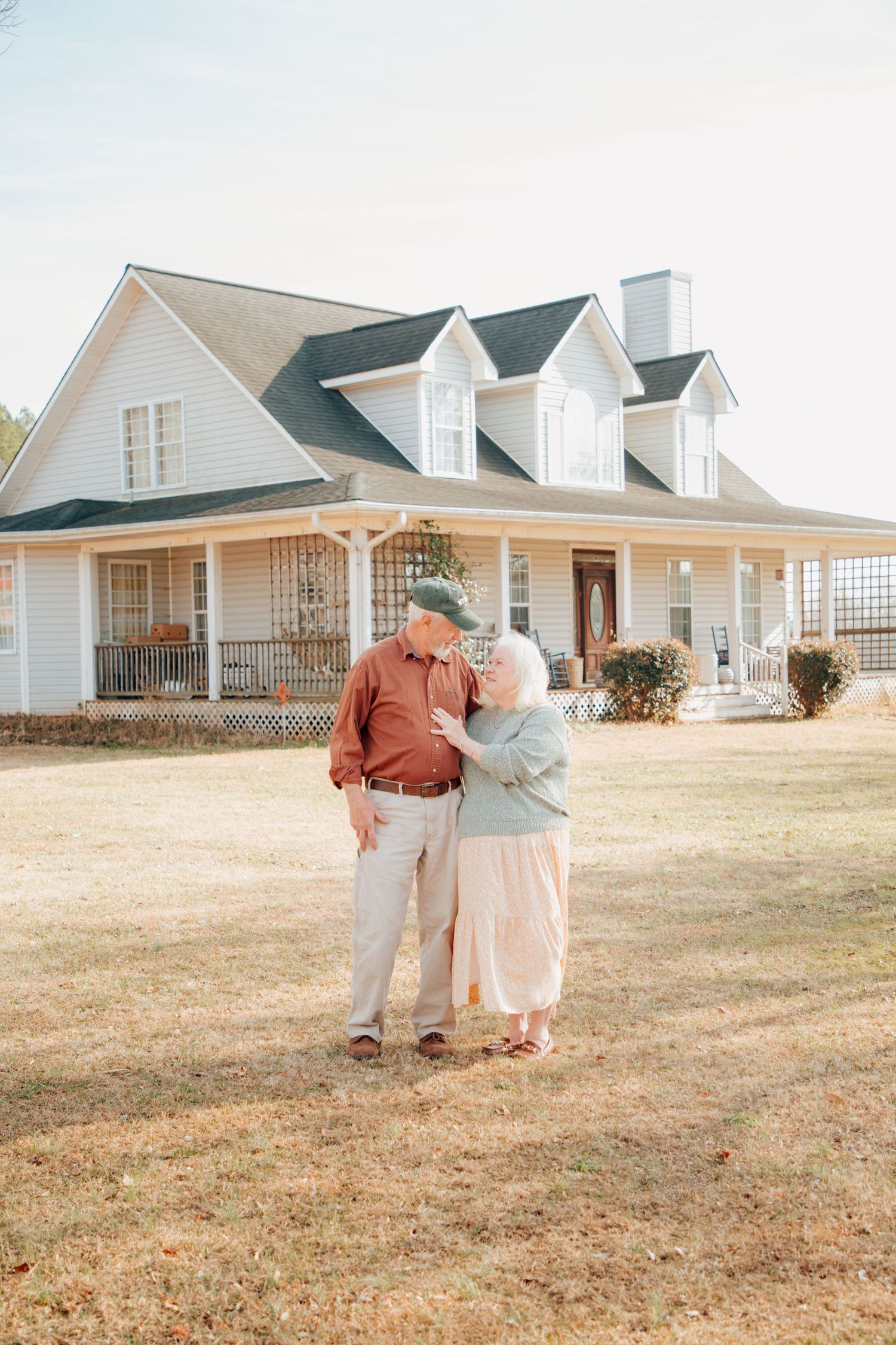 Elderly couple sharing a tender kiss in front of a white farmhouse on a sunny day.