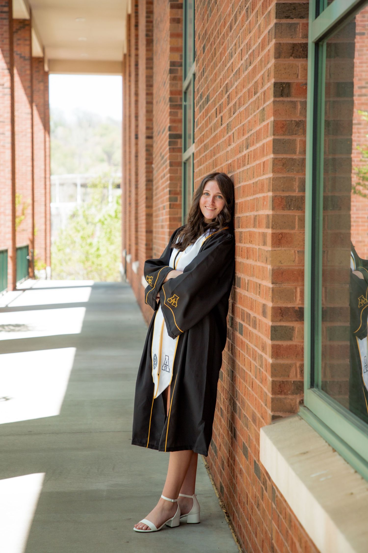 Smiling graduate in black robe leaning against brick wall in hallway.