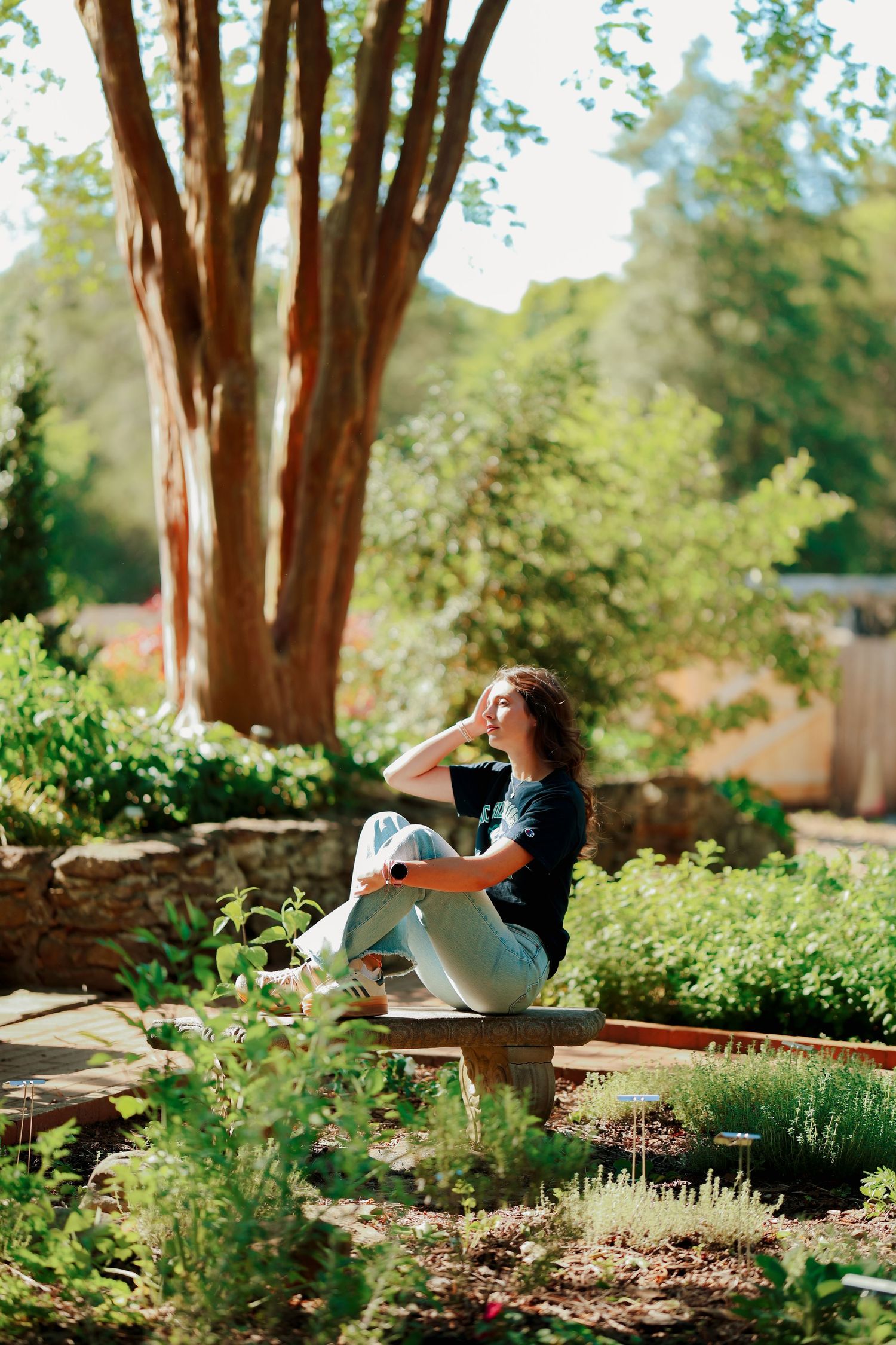 Young woman in dark shirt sitting on a garden bench surrounded by lush greenery and trees in sunlight.