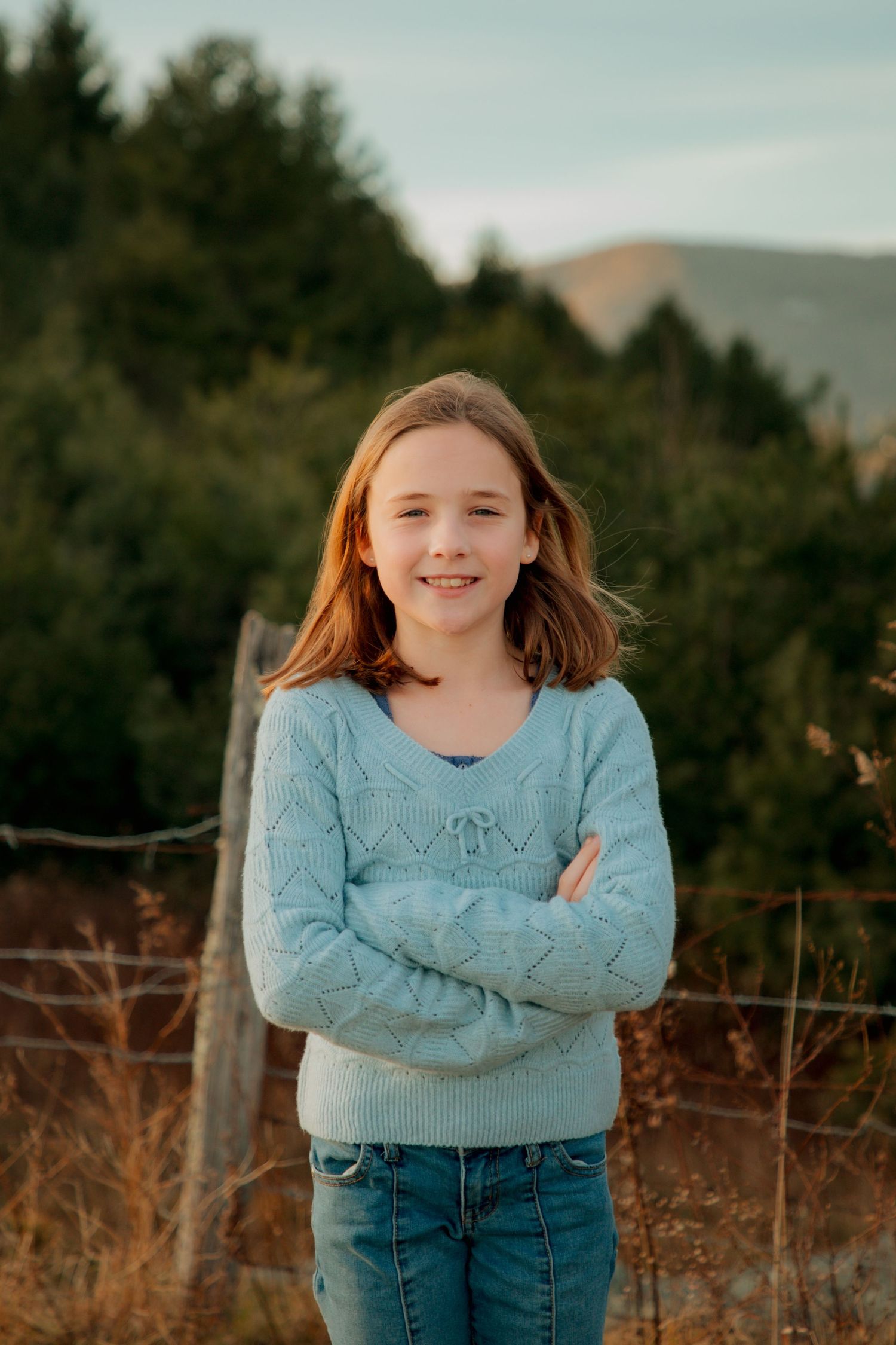 Smiling young girl with long hair crosses arms wearing light blue sweater and jeans with mountain backdrop.