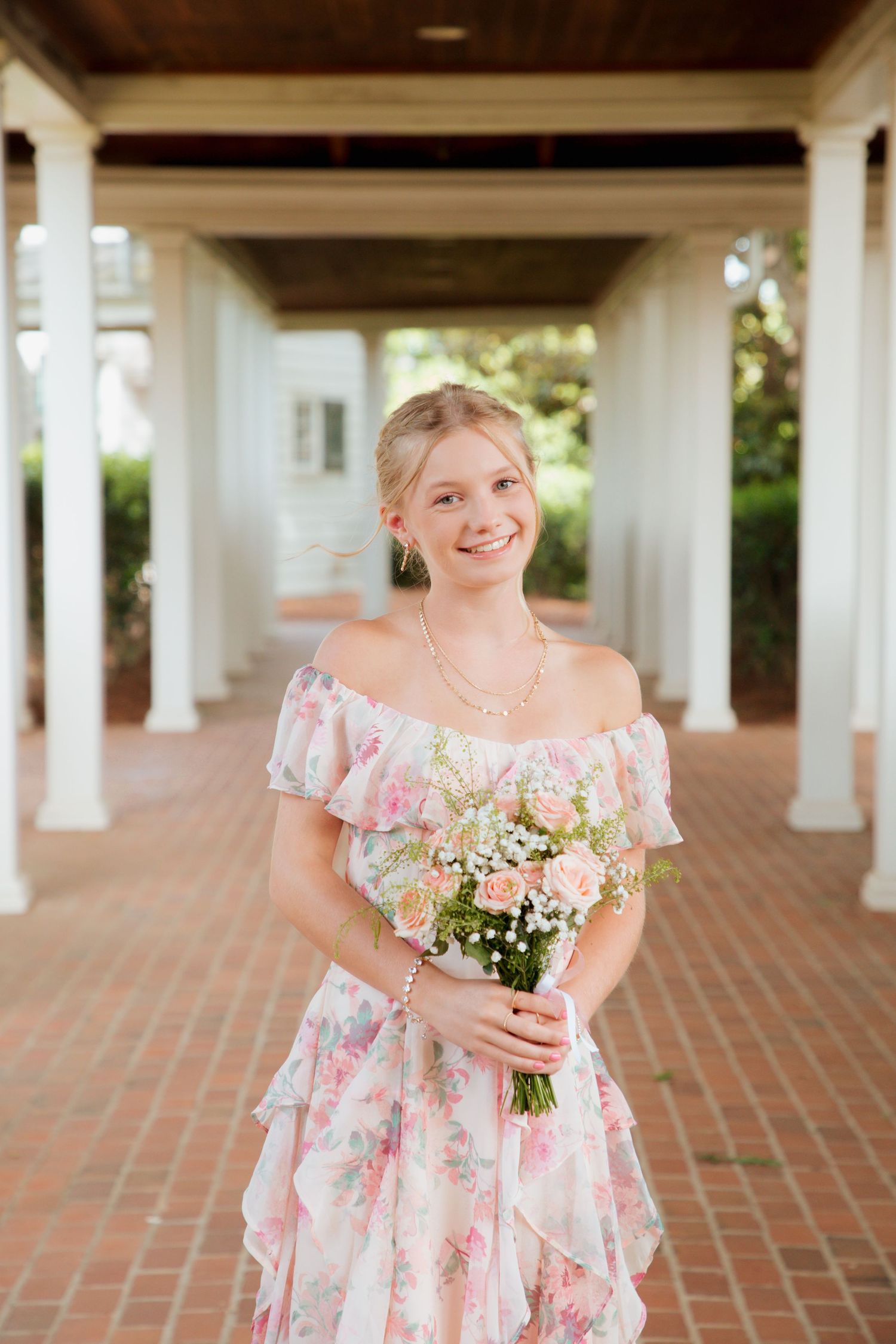 Smiling teen girl in off-shoulder floral dress holds bouquet under white columned pergola.