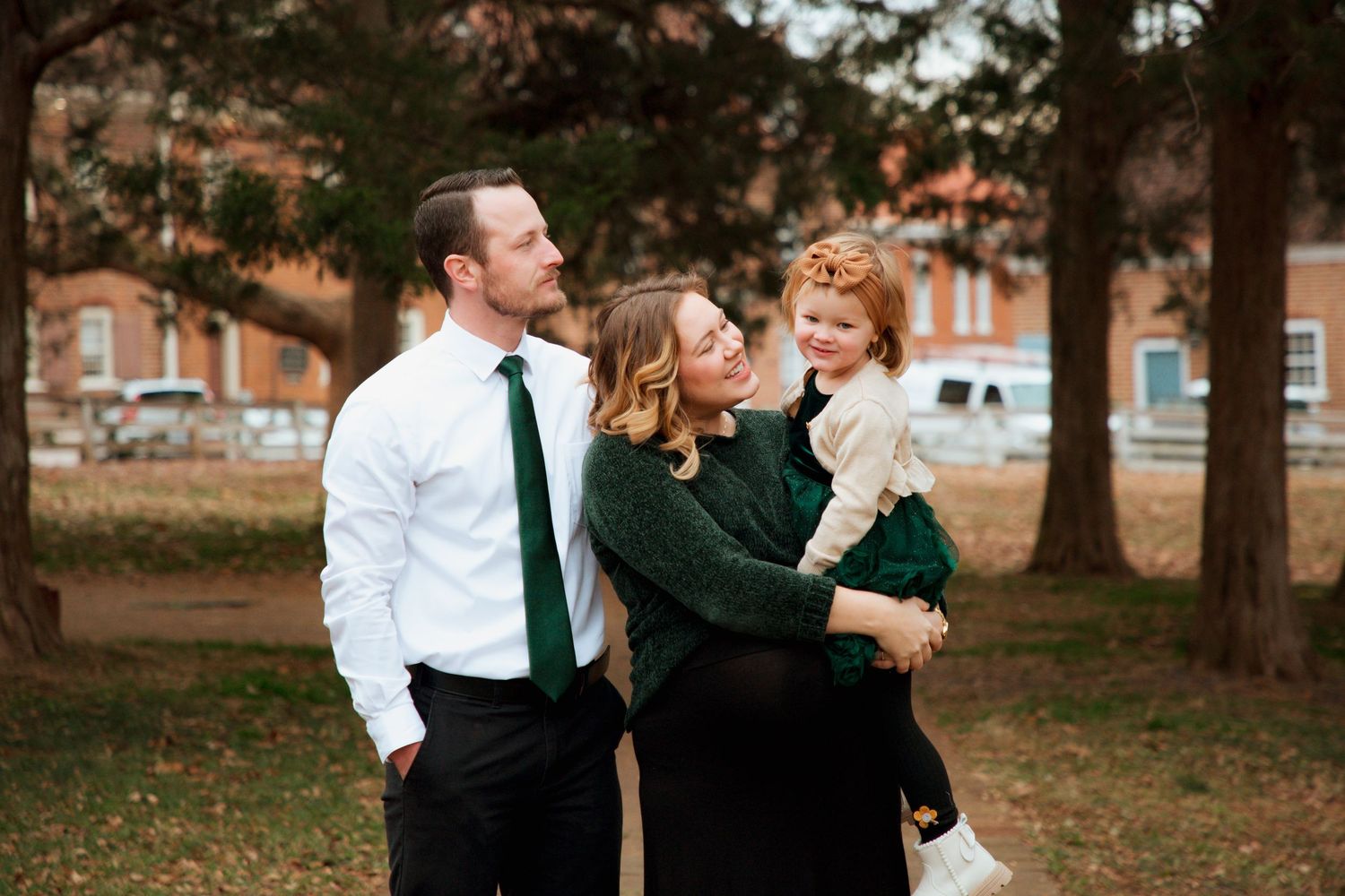 Family portrait of pregnant mother holding red-haired toddler with father in white shirt and green tie outdoors.