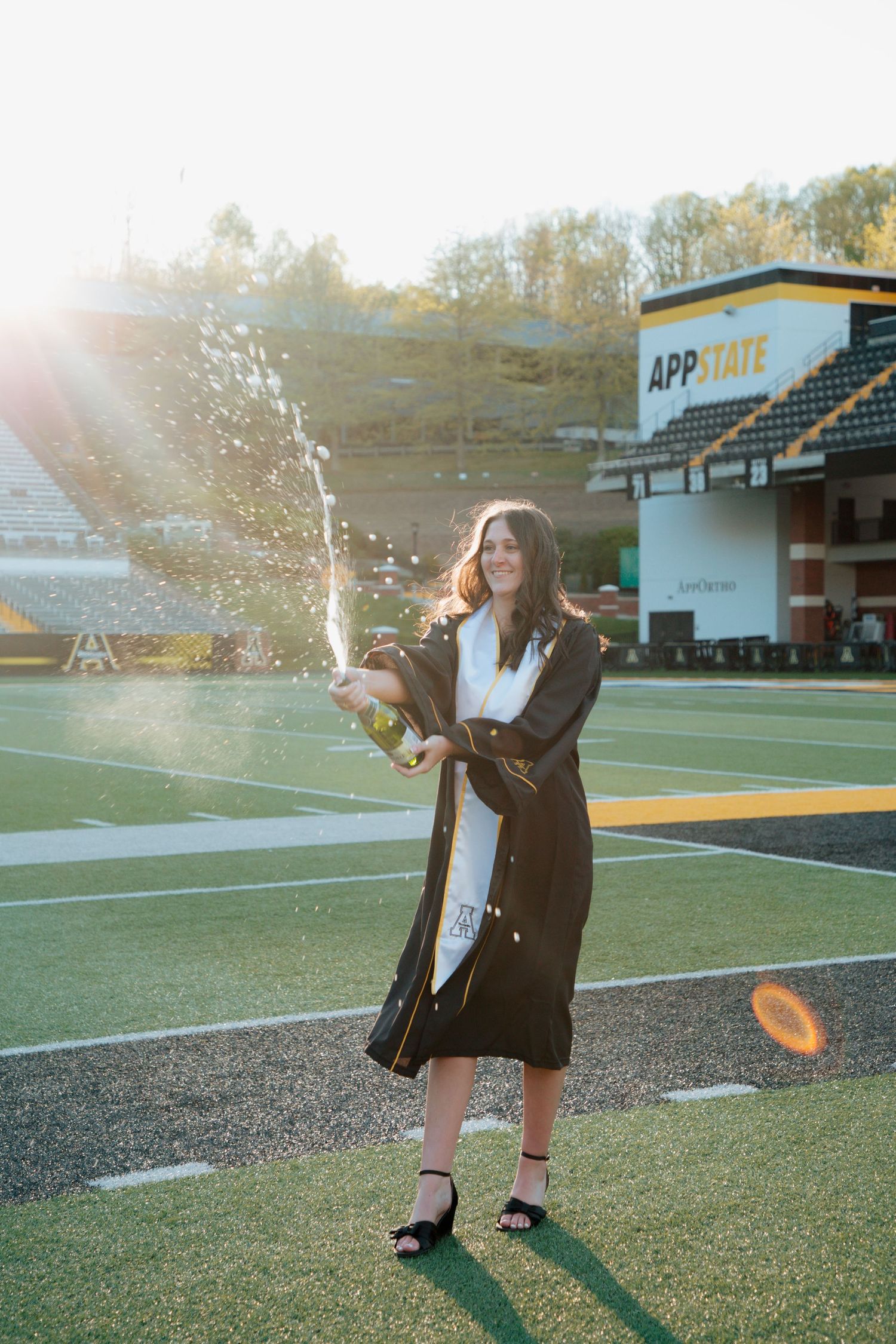 Graduate in black gown pops champagne bottle on App State football field at sunset.