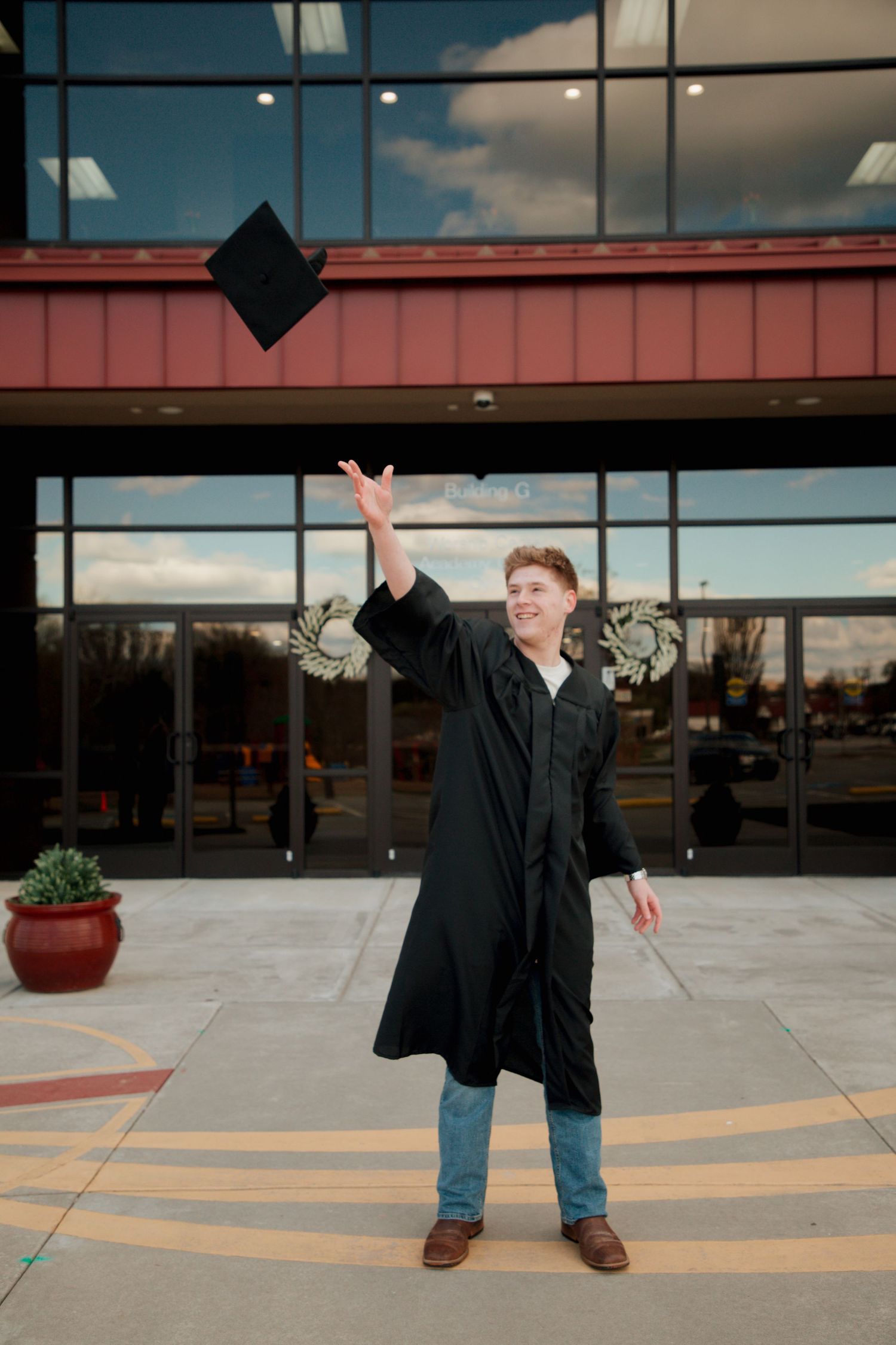 Graduate in black cap and gown tossing mortarboard in air outside modern building entrance.