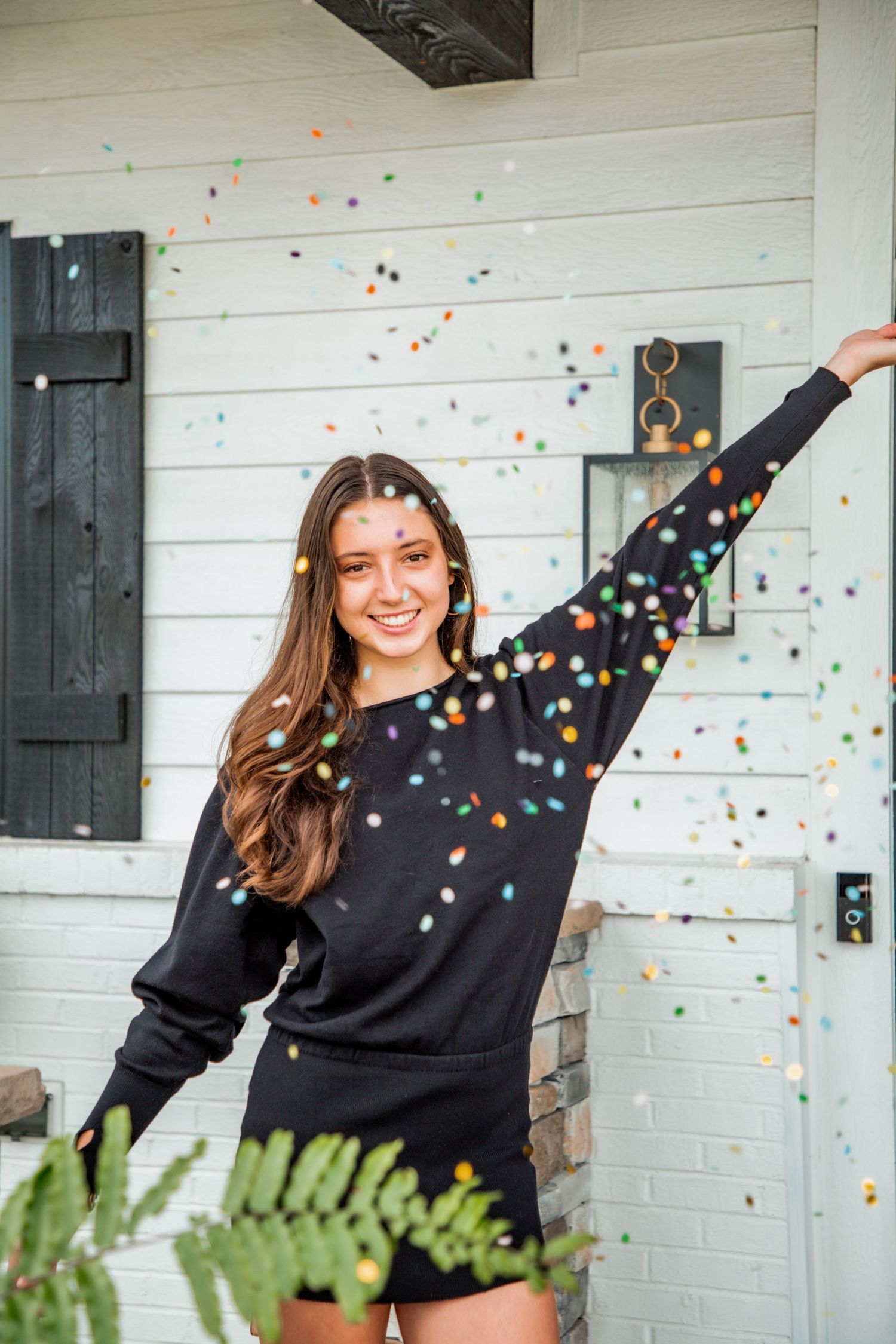 Joyful young woman in black sweatshirt celebrating with colorful confetti falling around her outdoors.