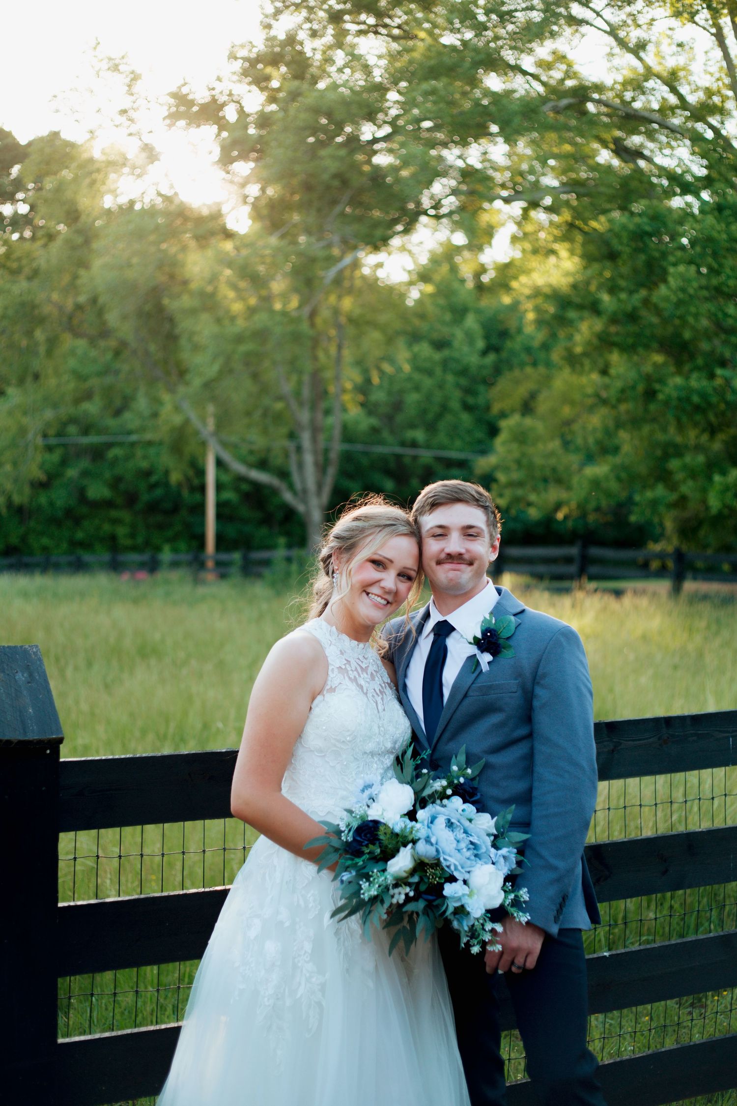 Smiling bride and groom pose by black fence holding blue and white bouquet.