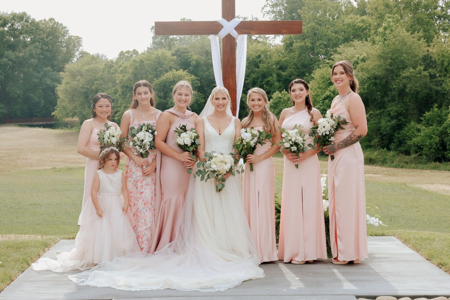 Bride with bridesmaids in blush pink dresses and flower girl holding white bouquets before wooden cross at outdoor ceremony.