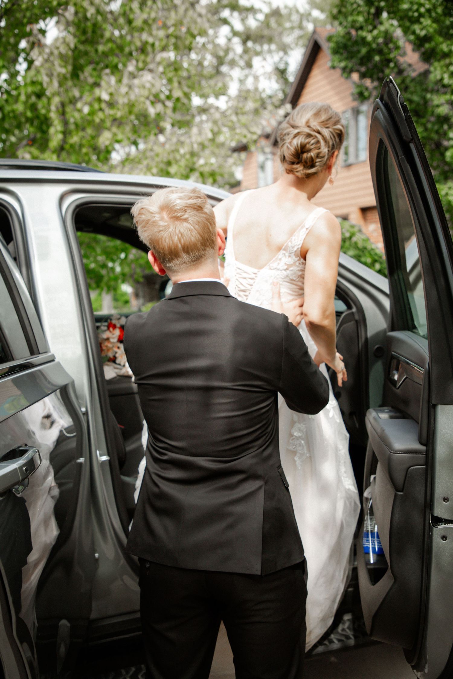 Groom in black suit assists bride in white lace wedding gown into a vehicle on their wedding day.