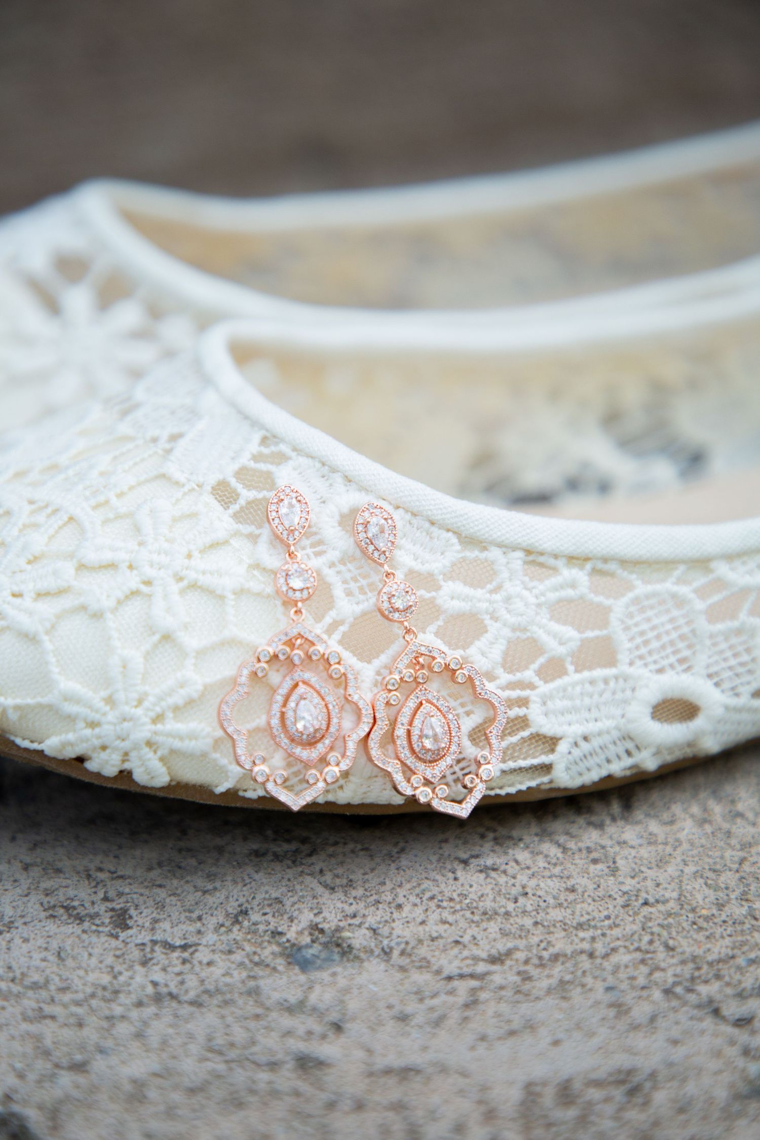 Elegant rose gold crystal drop earrings displayed on a white lace bridal flat shoe on stone surface.