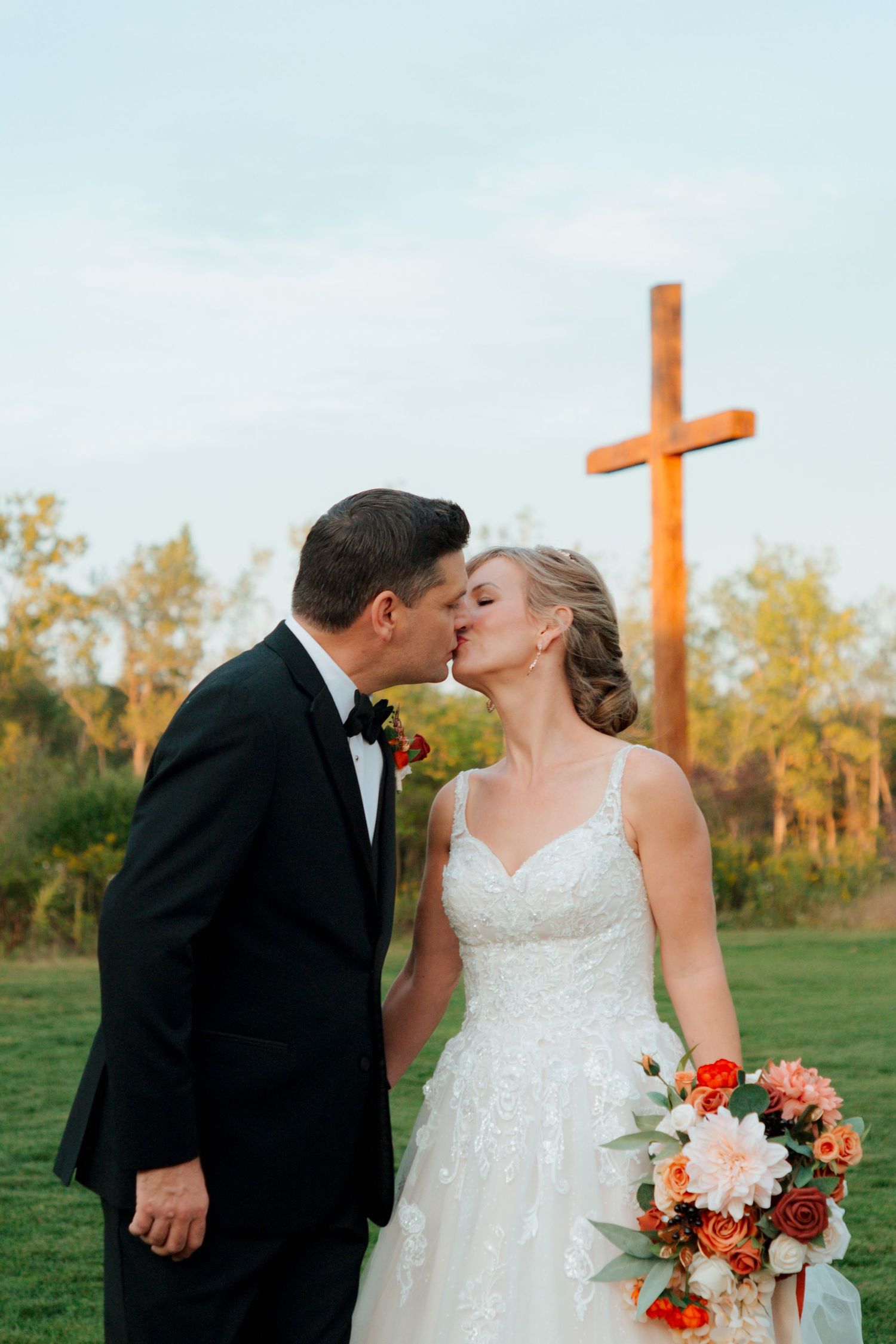 Bride and groom share a kiss before wooden cross, bride holding autumn floral bouquet.